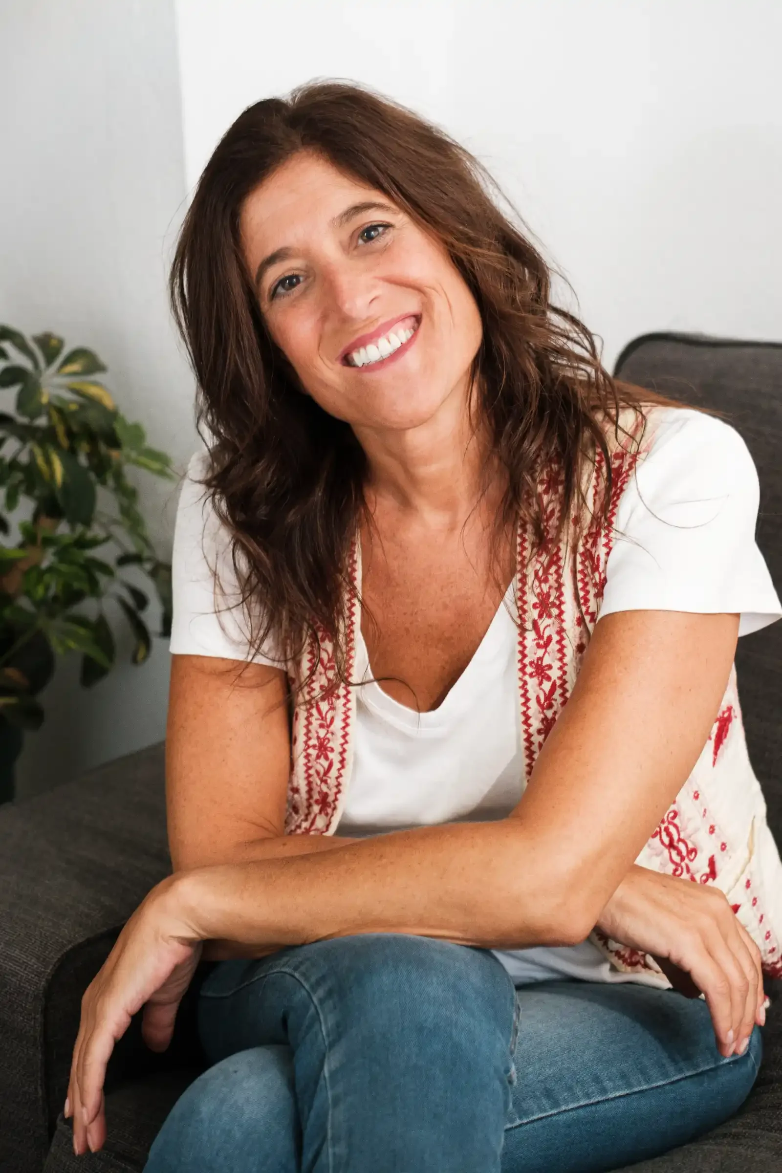 Retrato de una mujer sonriendo, con cabello castaño, camiseta blanca con detalles bordados en rojo y jeans azules, sentada en un sofá con una planta de fondo.