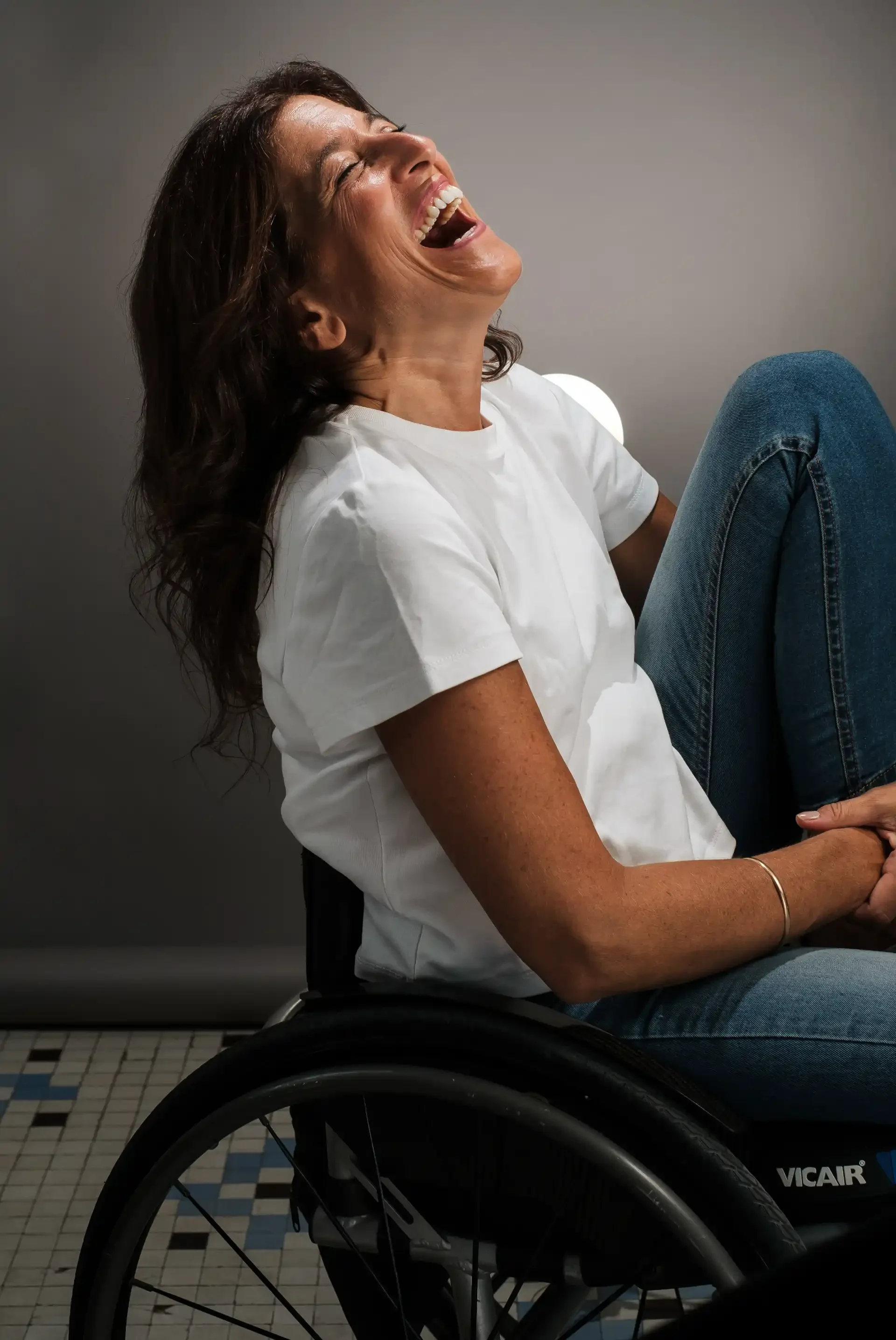 Mujer sonriente y riendo en silla de ruedas, usando camiseta blanca y jeans, en un fondo gris.