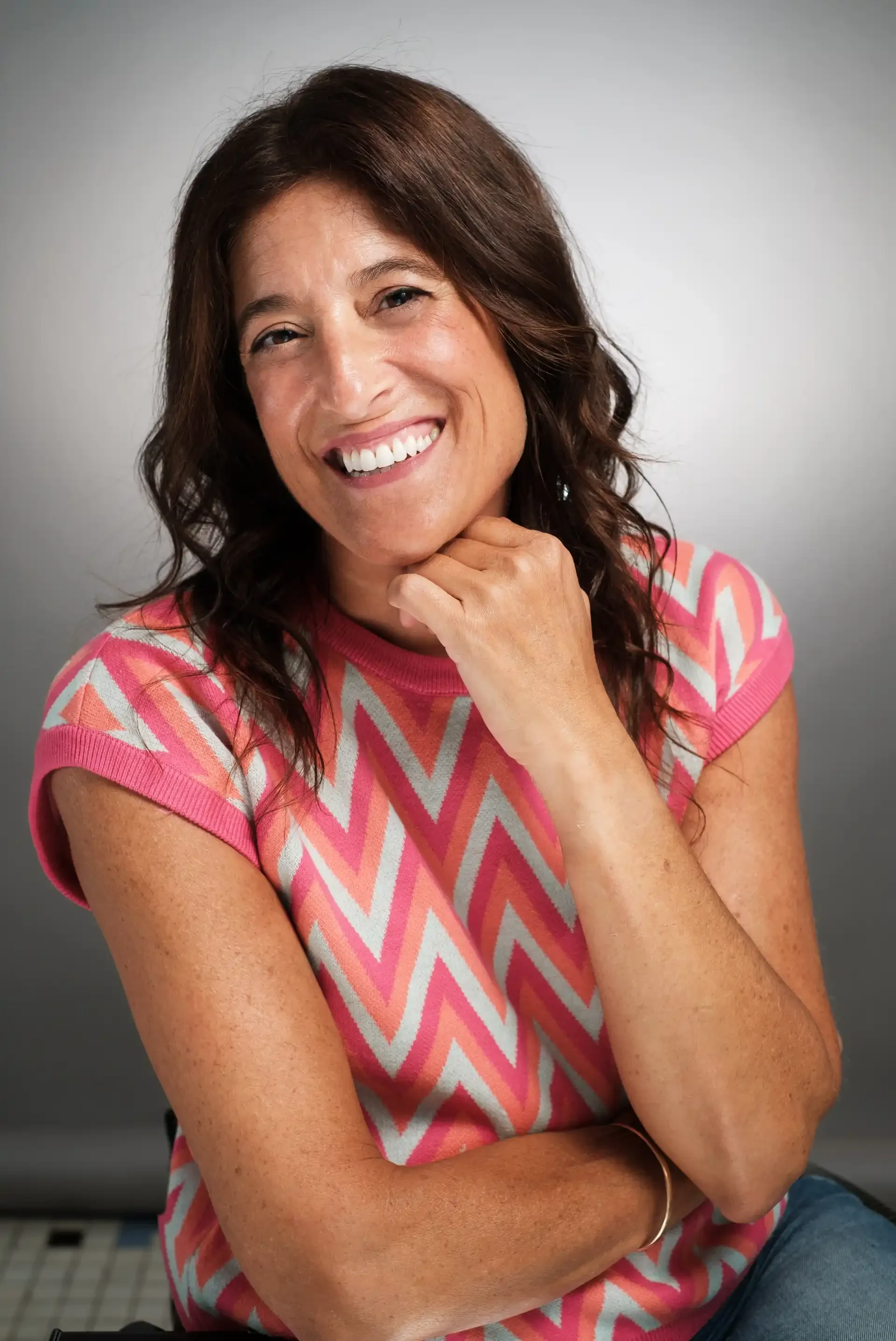Mujer con cabello castaño oscuro y ojos marrones, sonriendo y con la mano en el cuello, usando una camiseta de manga corta con patrón en zigzag en tonos rosas, beige y gris, frente a fondo gris.