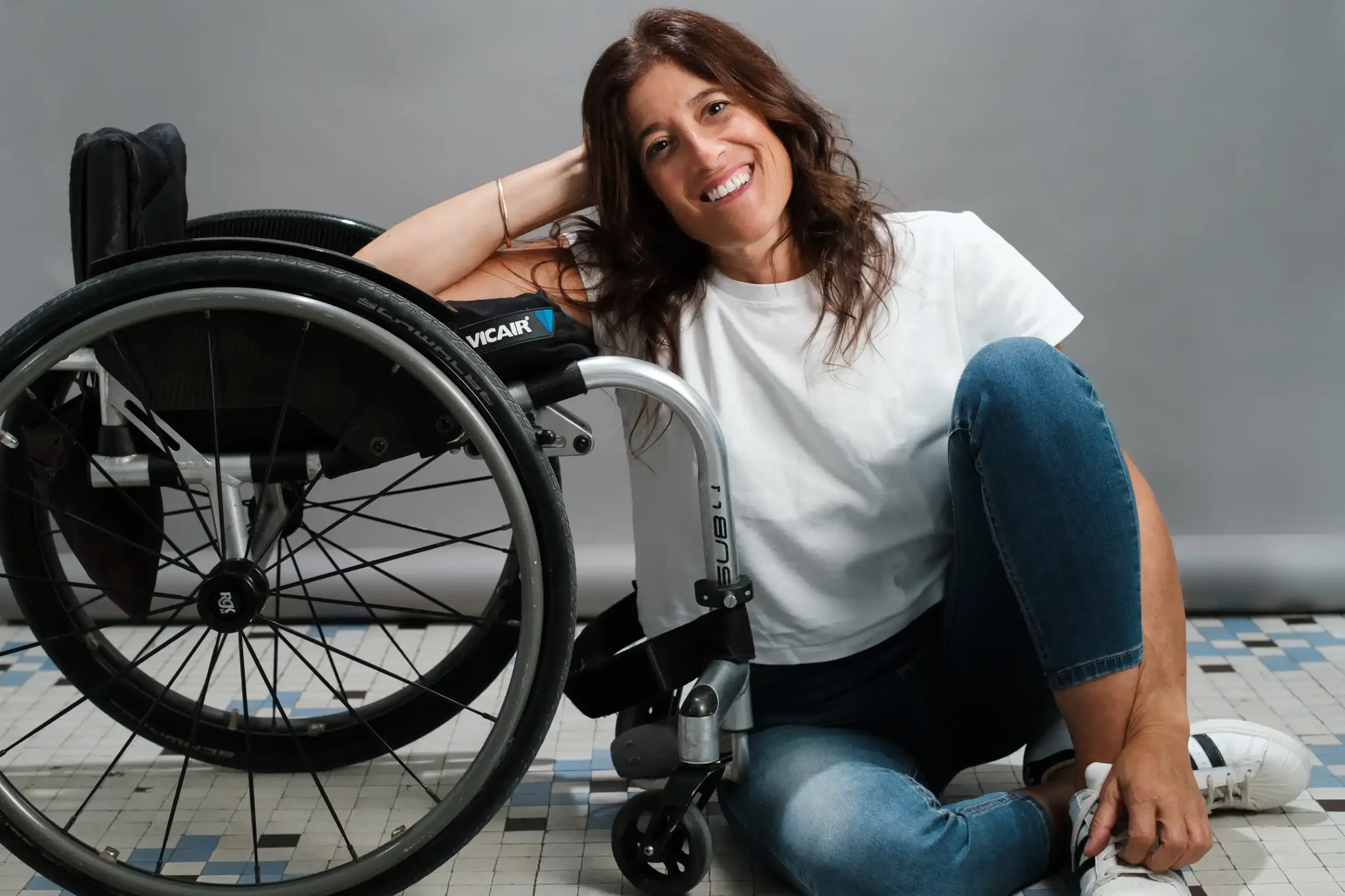 Mujer sonriente en una silla de ruedas, vestida con camiseta blanca y jeans azules, en un espacio con paredes grises y suelo de baldosas. La mujer está agachada, con una mano en su pierna y la otra apoyada en la cabeza.