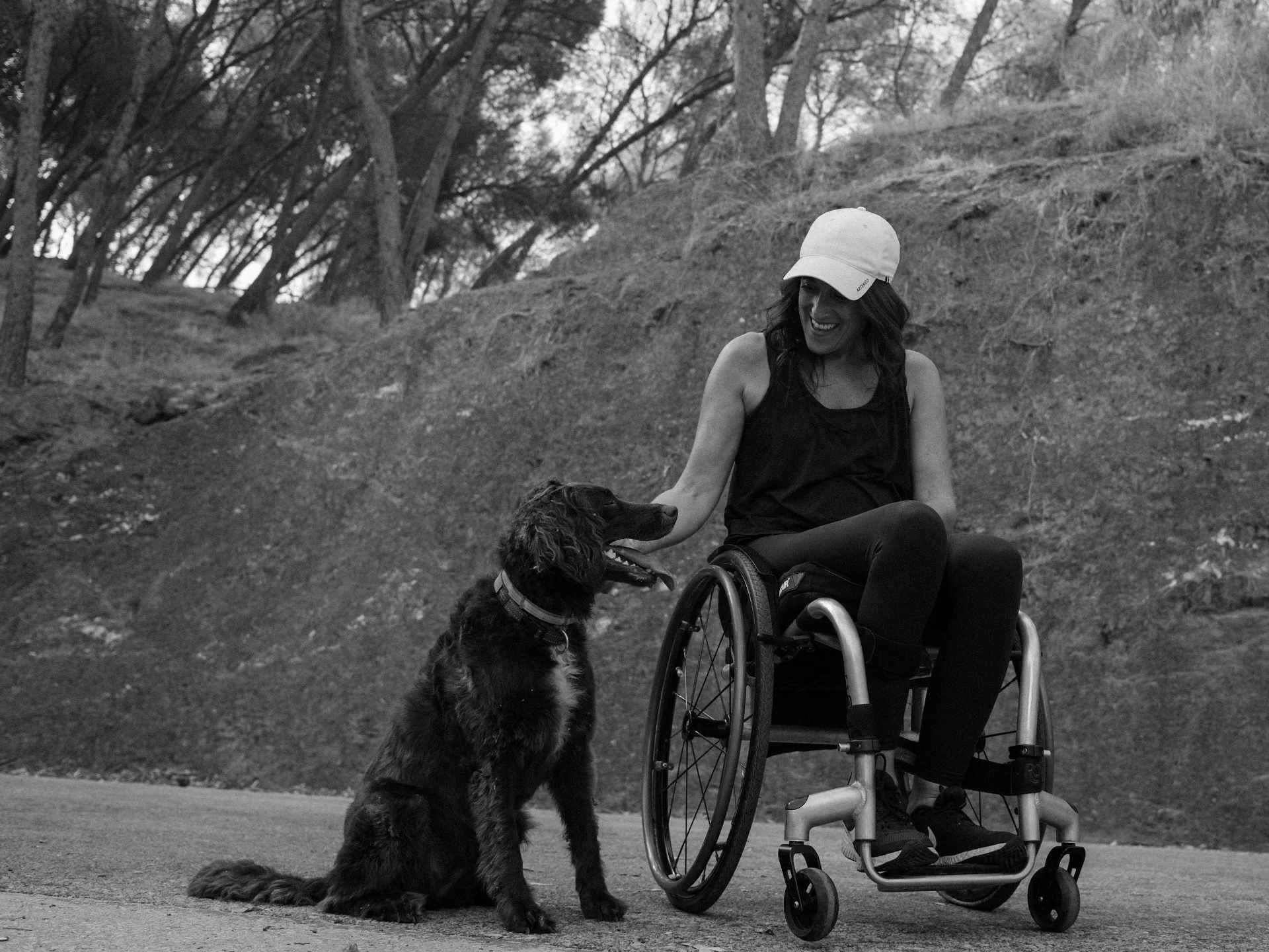 Mujer en silla de ruedas sonriendo y acariciando a un perro grande en un parque, en blanco y negro.