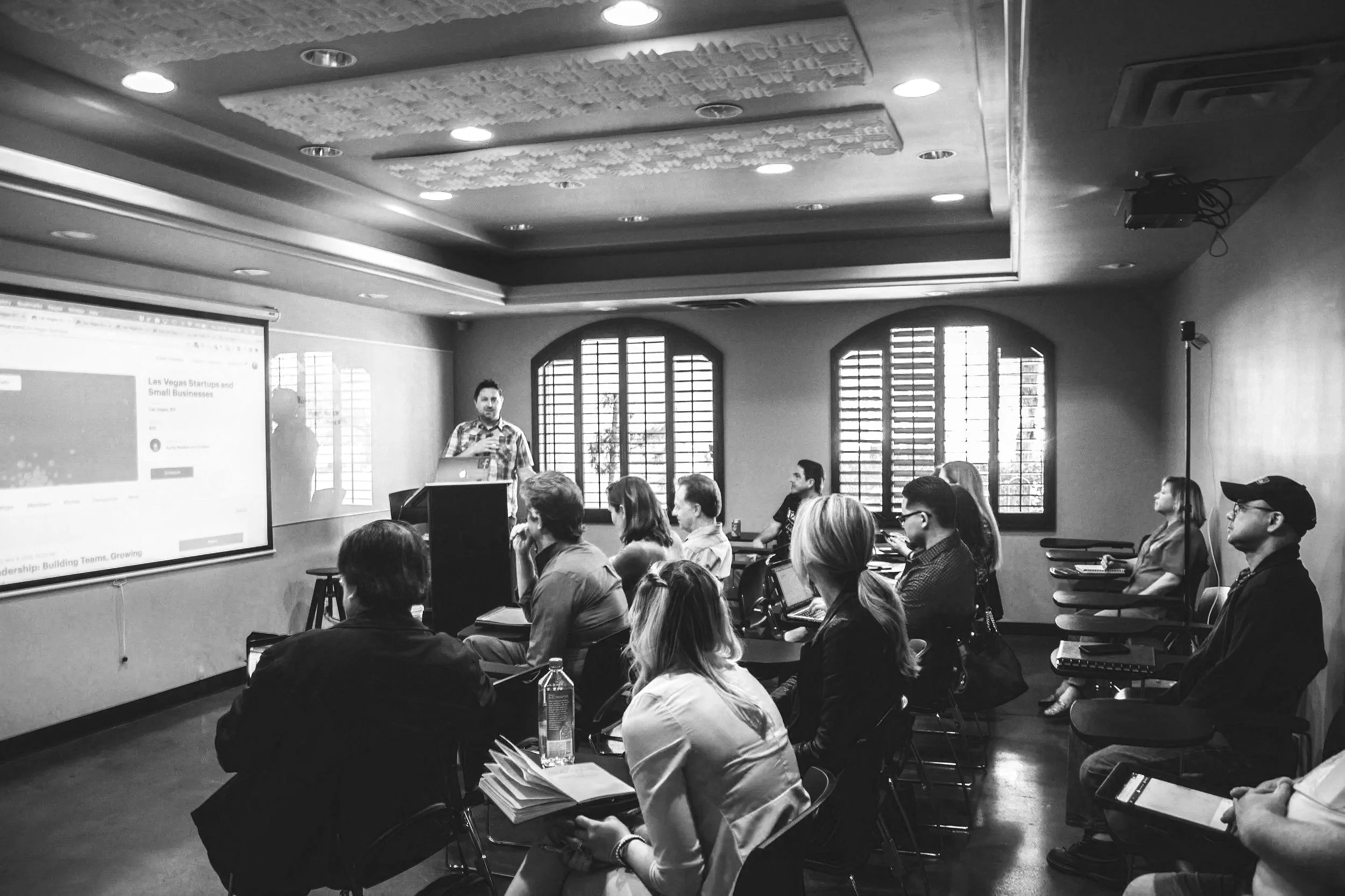 Reunión o presentación en una sala con asistentes sentados y un orador de pie frente a una pantalla, en una habitación con ventanas con barrotes y luz natural.