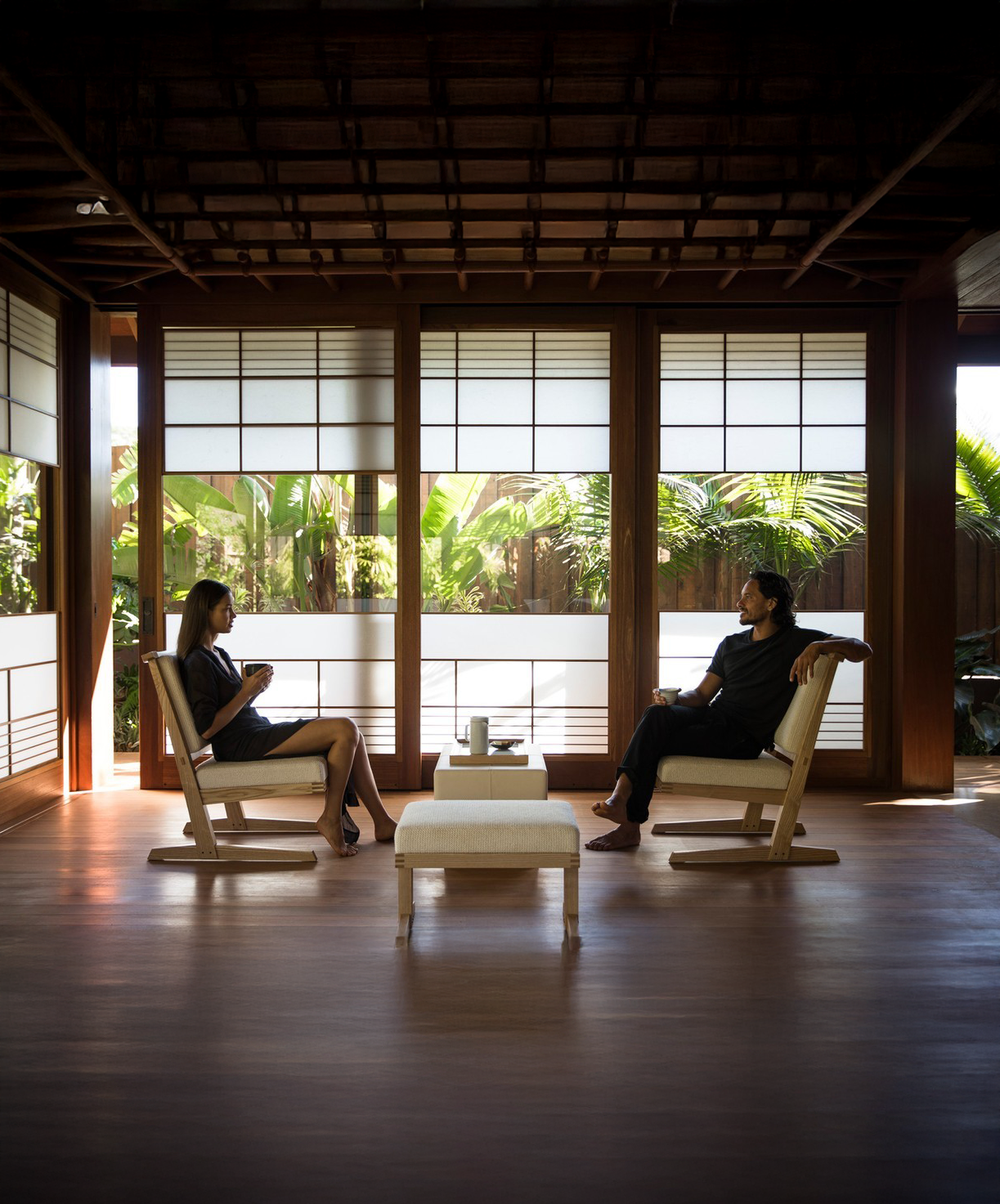 Two people sitting in a sunlit room with Japanese-style shoji screens, having a conversation while drinking tea.