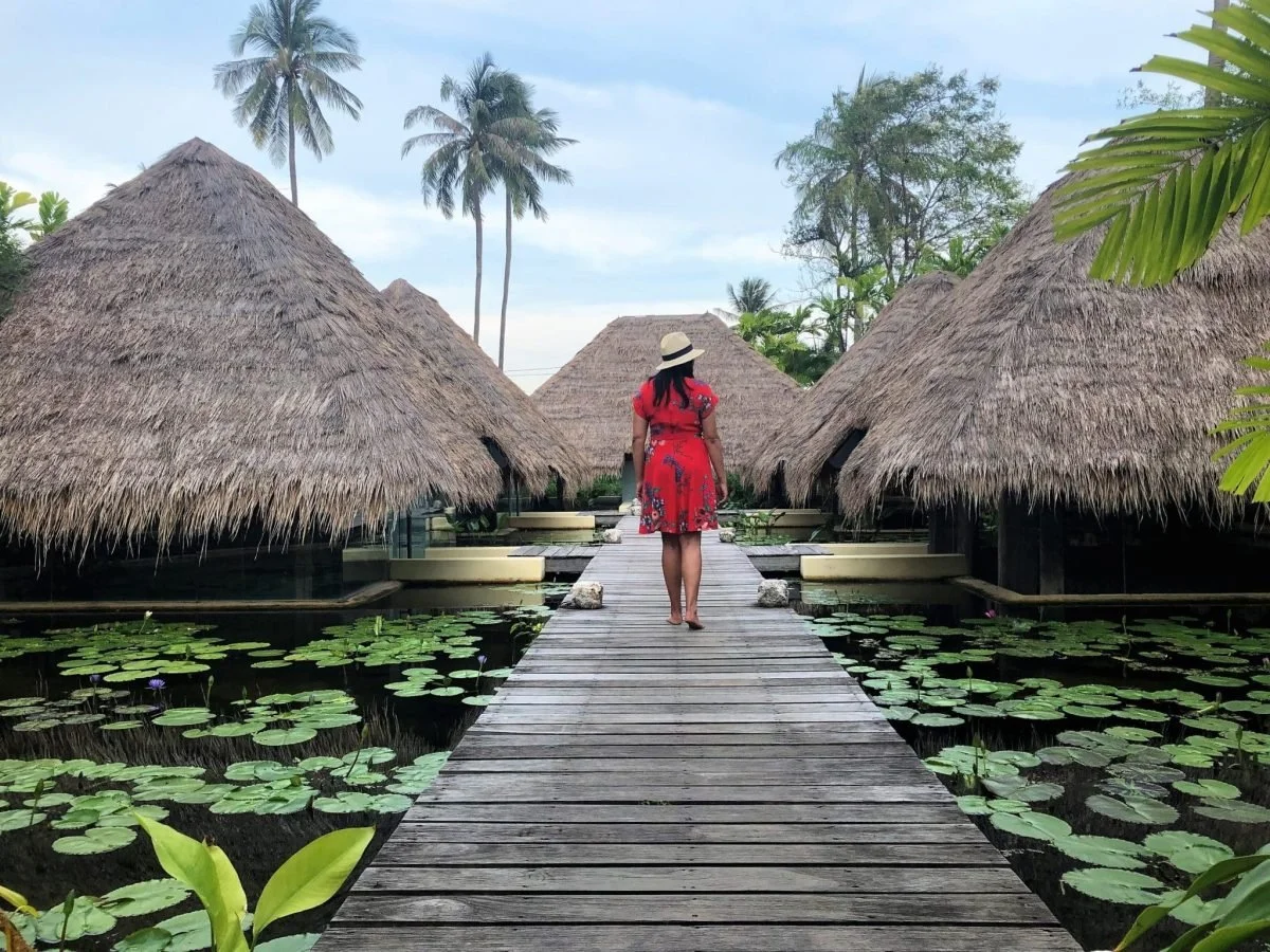 A woman in a red dress and straw hat walking on a wooden bridge over a pond with lily pads, surrounded by thatched-roof huts and tropical trees.