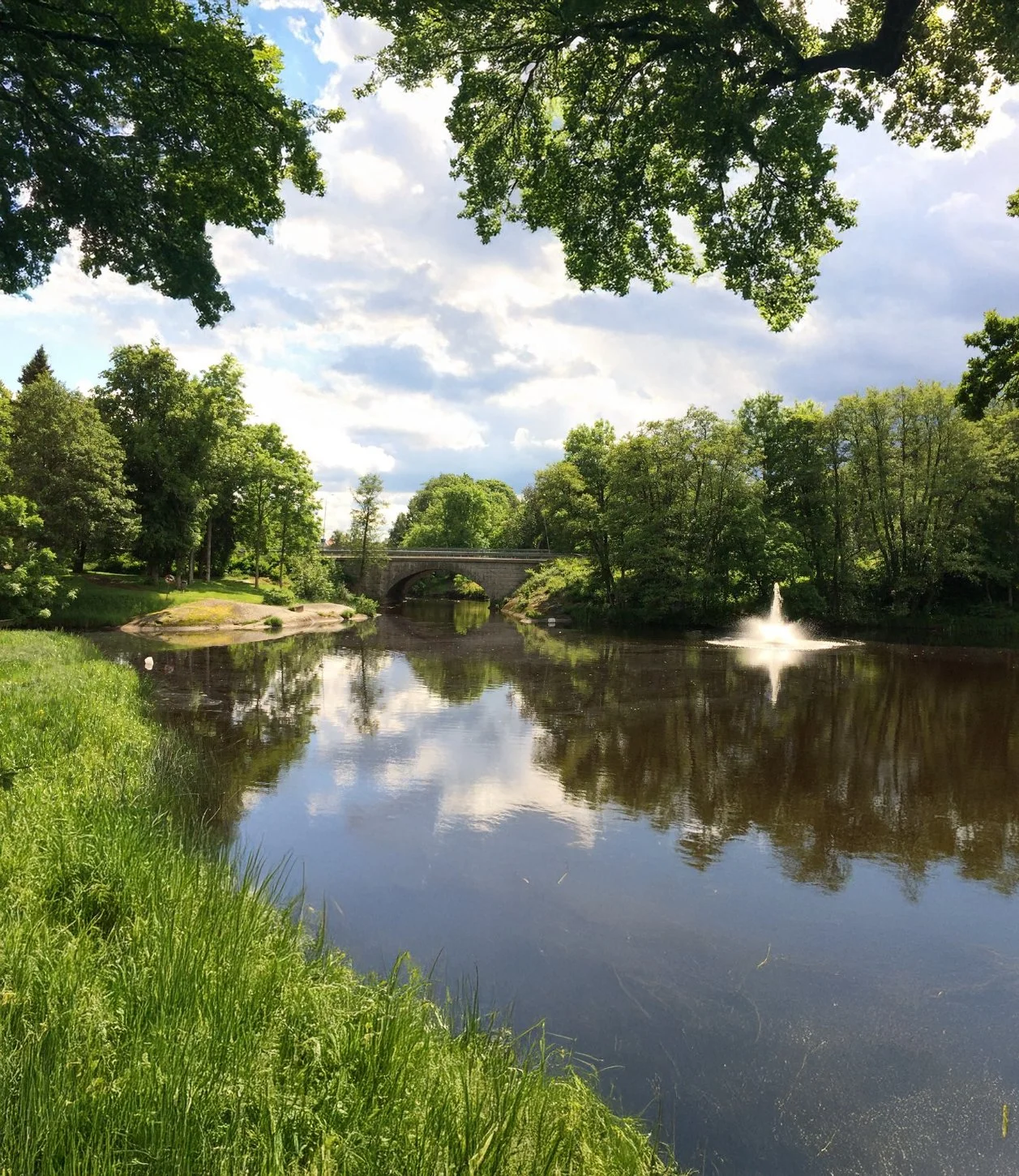 A scenic view of a river with a fountain on the right side, surrounded by green trees and grass, with a stone bridge in the background and a partly cloudy sky.