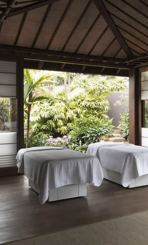 Two massage tables covered with white sheets in a cozy room with open windows showing lush green plants outside.