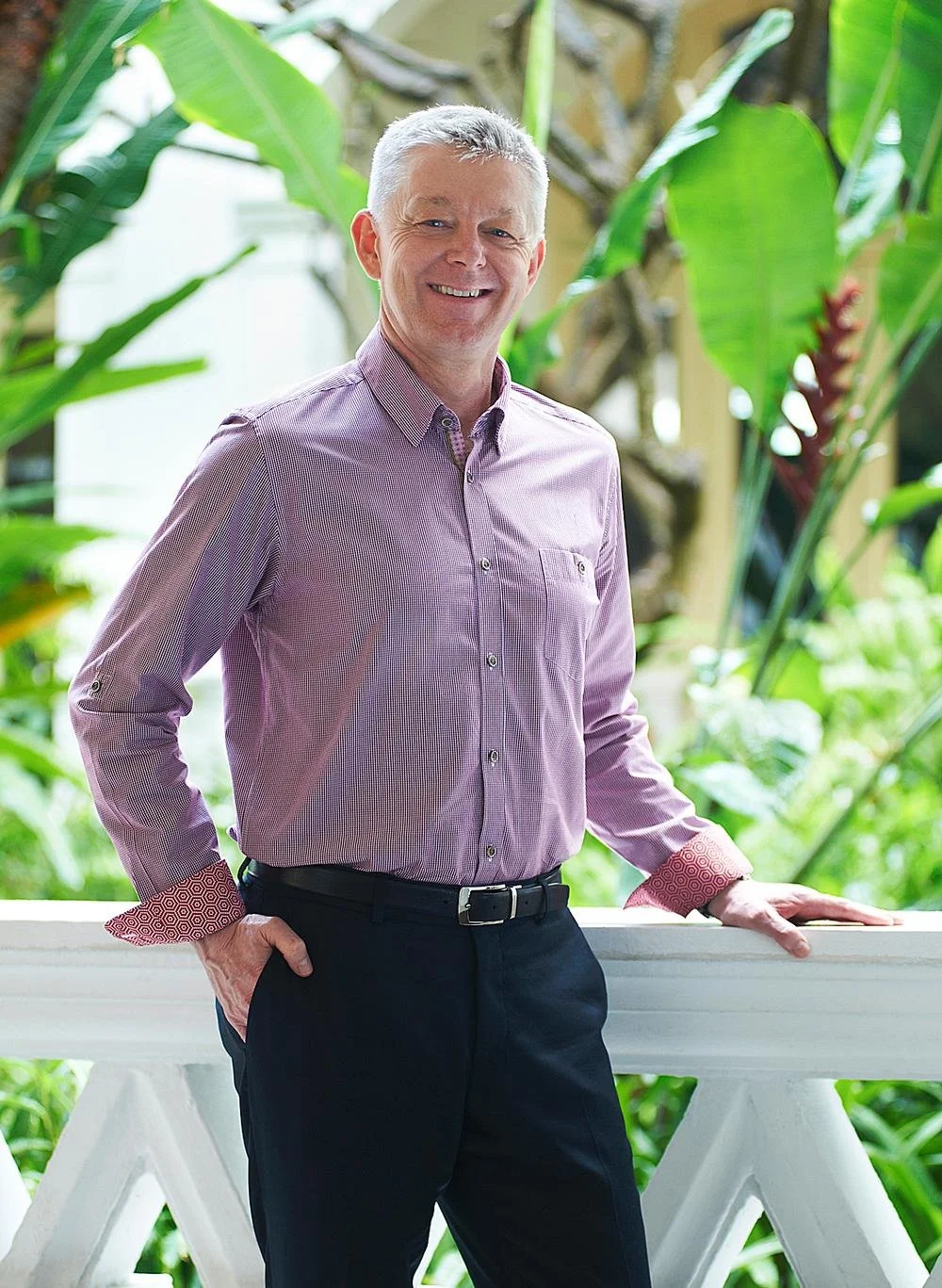 A middle-aged man with gray hair, wearing a light purple button-up shirt with red patterned cuffs, standing outdoors with large green tropical leaves in the background. He is smiling and leaning on a white railing.