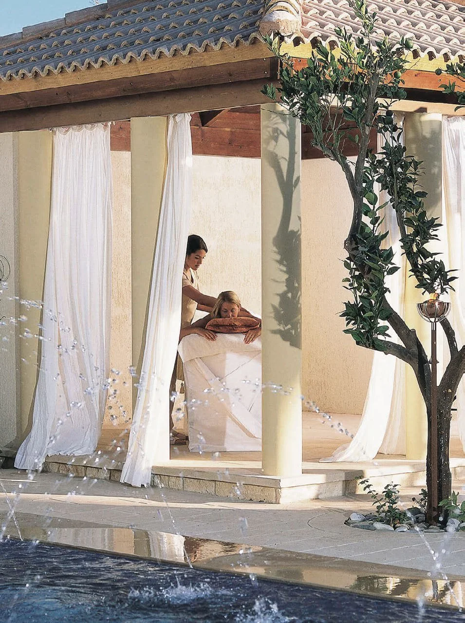 A woman receives a massage outdoors under a canopy with white curtains, next to a small tree and a fountain in the foreground.