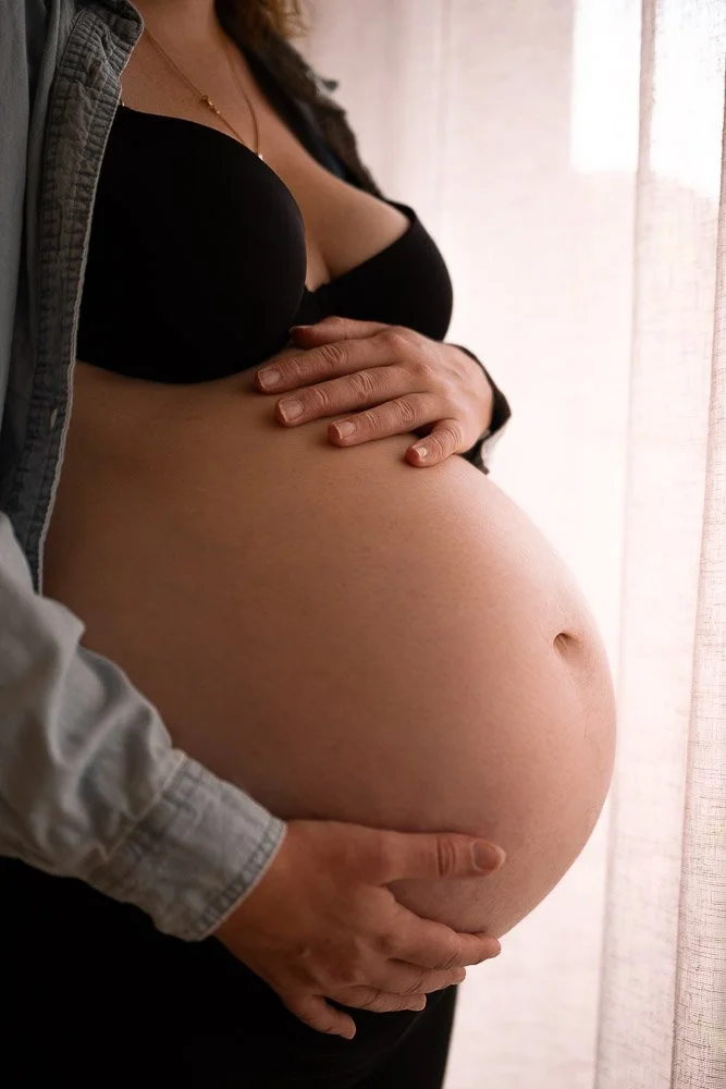 Femme enceinte tenant son ventre rond, portant un soutien-gorge noir et une chemise grise ouverte, près d'une fenêtre avec des rideaux Reportage photo bébé côte d’opale, Dunkerque, Calais, Wissant, Wimereux, Boulogne-sur-Mer, Hardelot-plage, Le Touqu