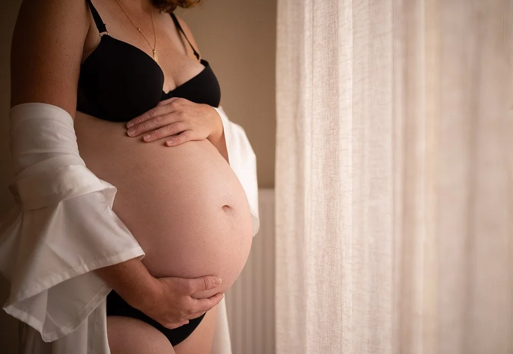 Une femme enceinte portant un soutien-gorge noir, une robe blanche décontractée, tenant son ventre avec ses mains, près d'une fenêtre avec des rideaux en tissu léger. Reportage photo grossesse intimiste côte d’opale, Dunkerque, Calais, Wissant, Wimer
