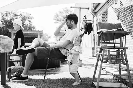 Un homme assis dans un fauteuil dehors, une petite fille lui tient la main, une autre personne marche au loin. Des meubles et des vêtements sont visibles à l'extérieur.