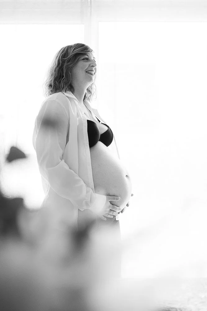 Femme enceinte souriante portant un soutien-gorge noir et une chemise blanche, debout près d'une fenêtre lumineuse.