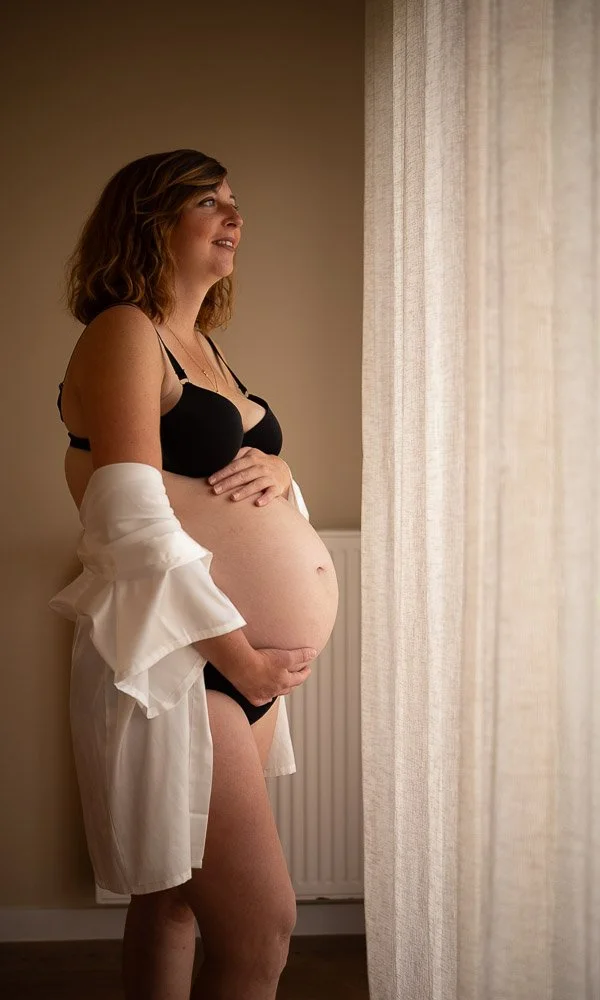 Une femme enceinte en sous-vêtements noirs et une chemise blanche ouverte, regardant par la fenêtre près d’un rideau léger. Reportage photo grossesse intimiste côte d’opale, Dunkerque, Calais, Wissant, Wimereux, Boulogne-sur-Mer, Hardelot-plage, Le T