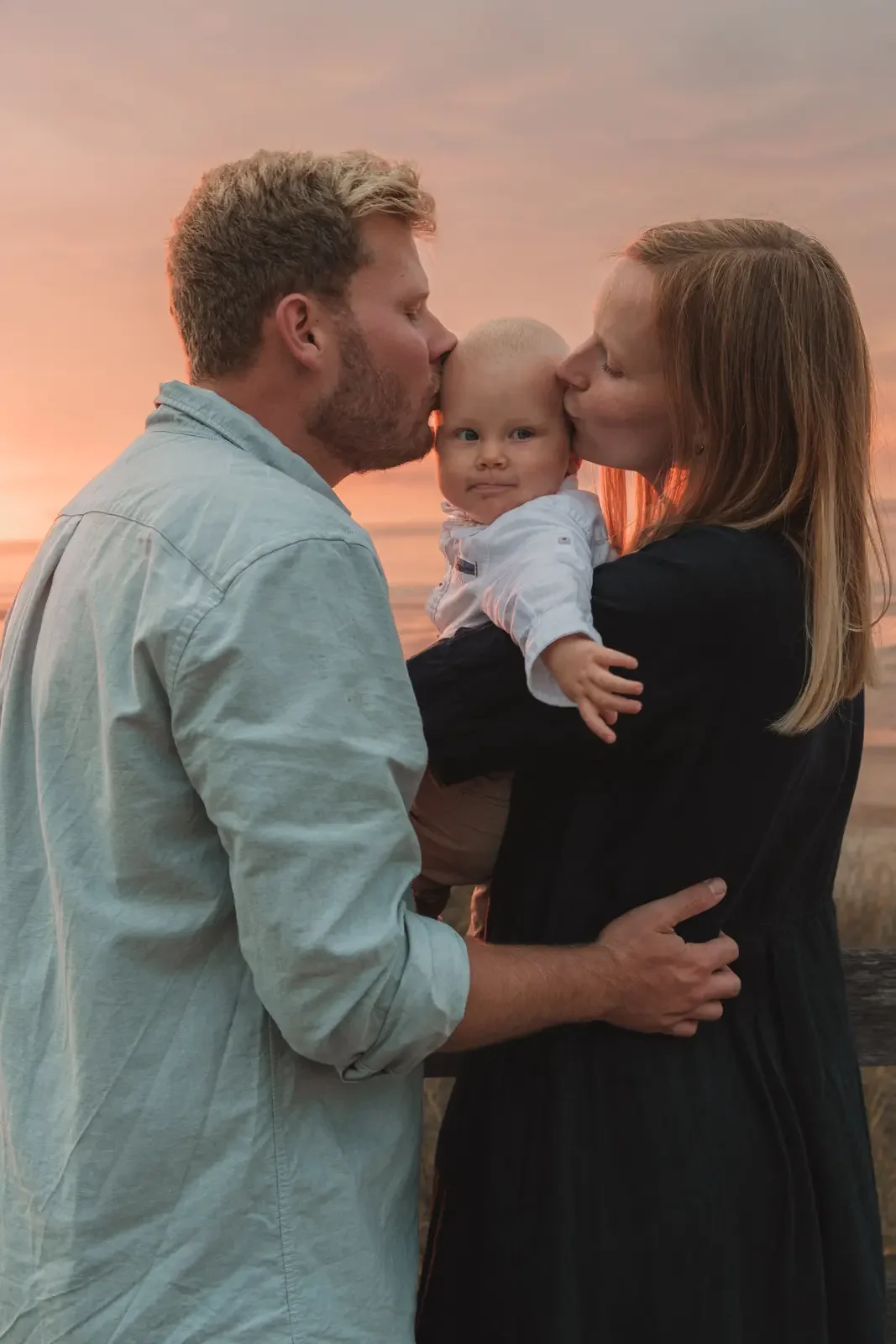 Une famille de trois personnes au coucher de soleil, un homme et une femme embrassant un bébé au centre. seance photo famille a domicile à wissant
