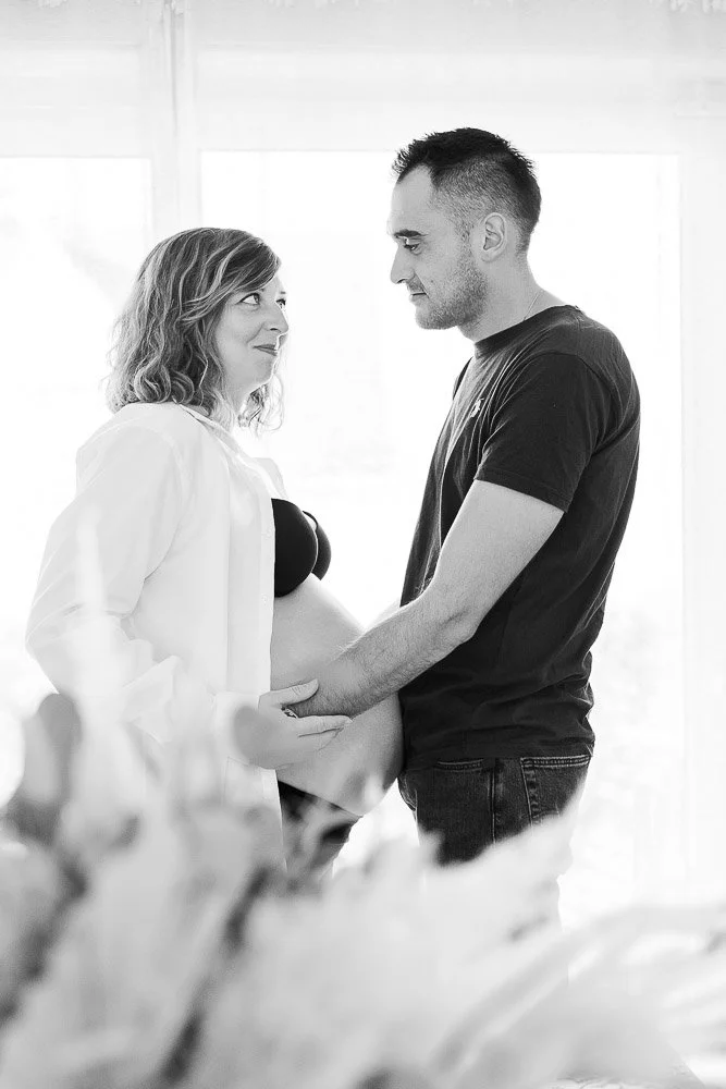 Une femme enceinte et un homme se tiennent face à face, se tenant la main, dans une pièce lumineuse. Reportage photo bébé côte d’opale, Dunkerque, Calais, Wissant, Wimereux, Boulogne-sur-Mer, Hardelot-plage, Le Touquet, Lille, sangatte.