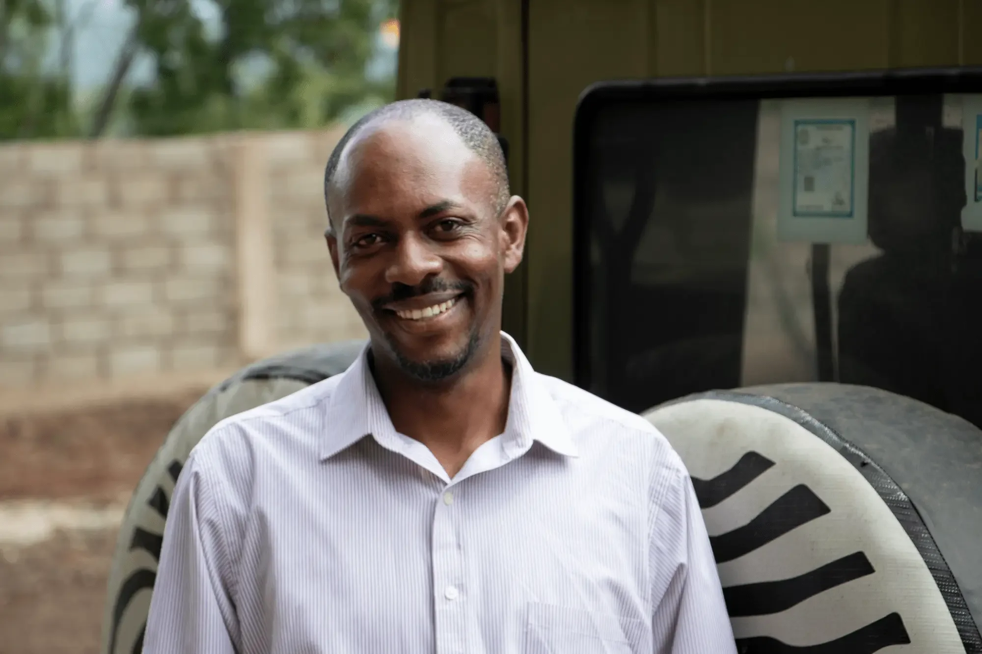 A smiling man standing outdoors near a large tire and a vehicle. He is wearing a white dress shirt with a collar.
