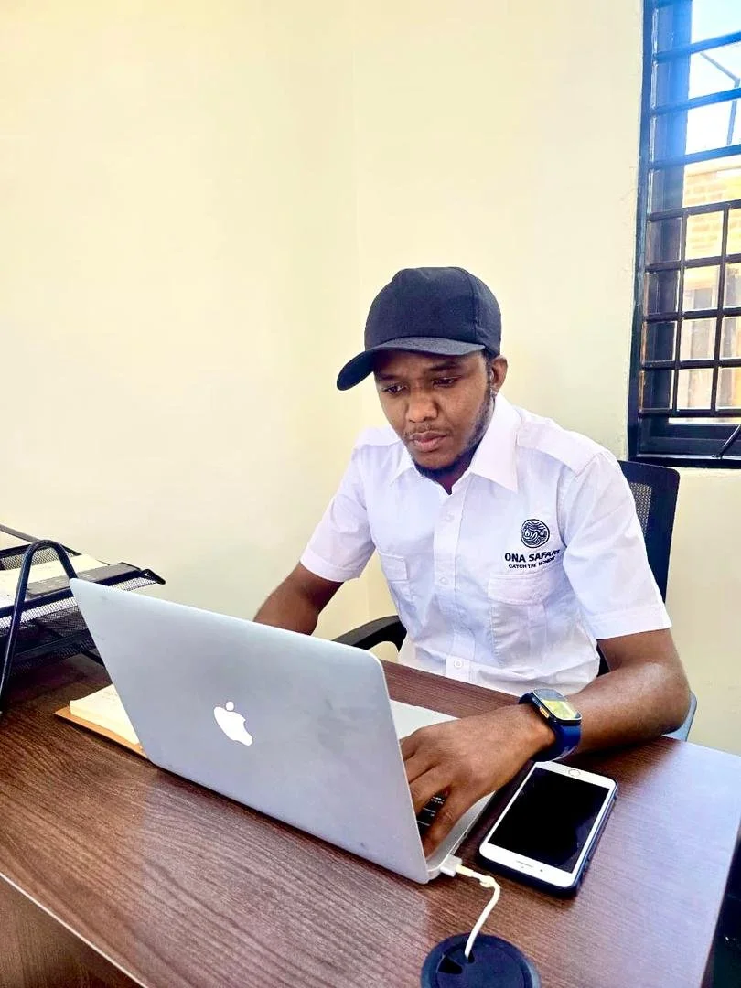 A man sitting at a desk using a silver MacBook laptop, wearing a white shirt, black cap, smartwatch, and with a smartphone connected via charging cable placed on the desk, in front of a window with metal bars.
