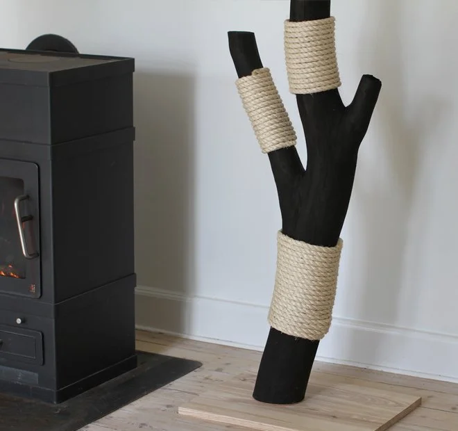 Black wooden cat scratching post with beige sisal rope, placed near a wood-burning stove