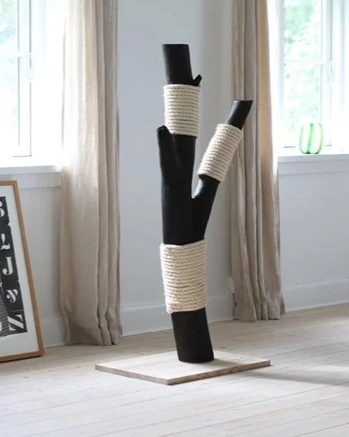 Sculptural black wooden cat scratching post wrapped with white rope, positioned near a window
