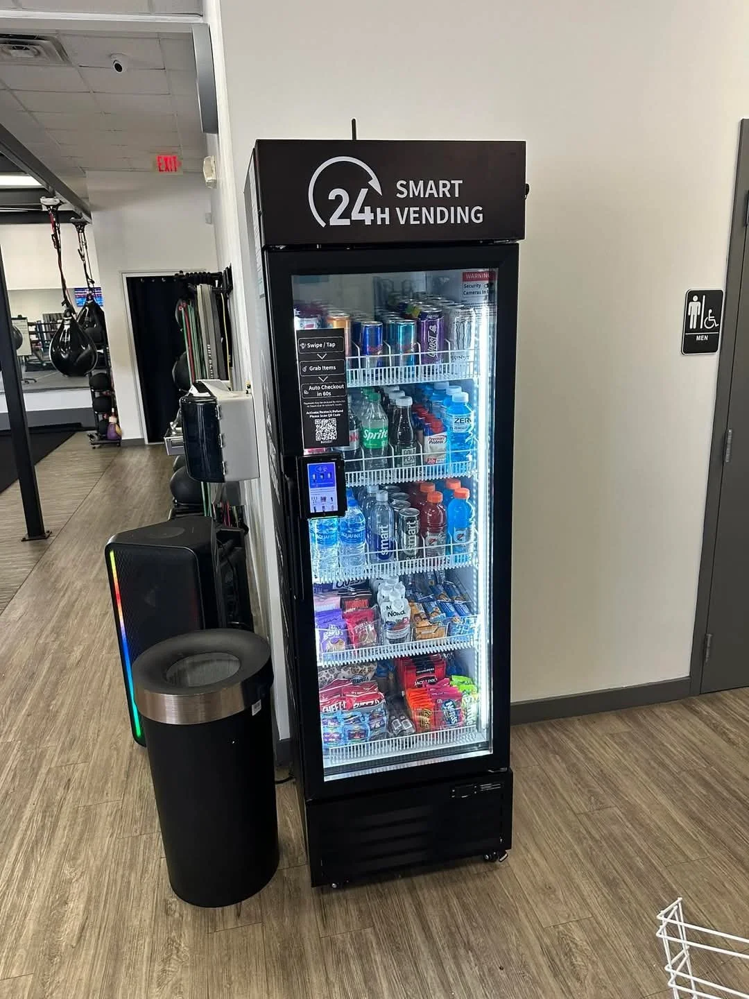A 24-hour vending machine stocked with sodas, water, and snacks, situated next to a trash can and a white wall, in a gym or fitness center environment.