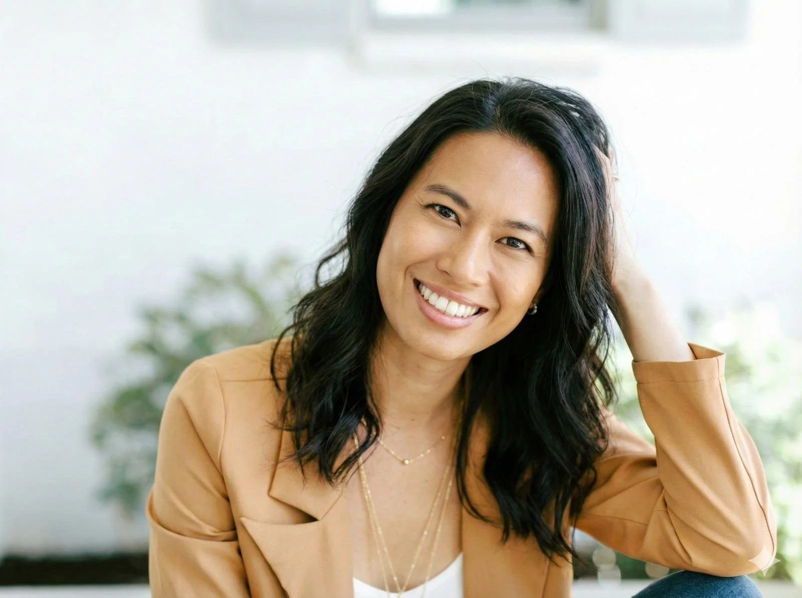 A closeup woman in a tan blazer resting her head against her hand, smiling at the camera.