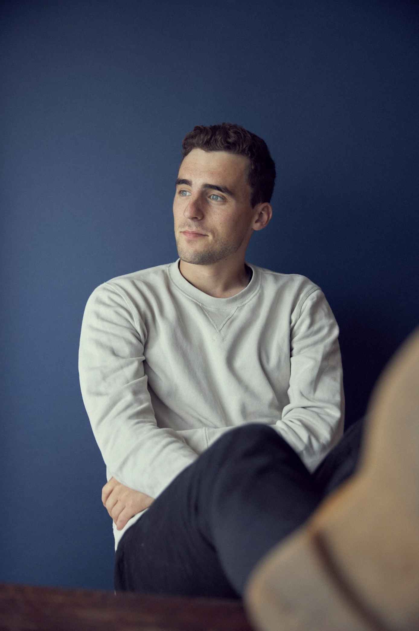 A young man with short, dark hair and light skin, wearing a light gray sweatshirt, sitting with his arms crossed against a solid dark blue background, looking to the side.
