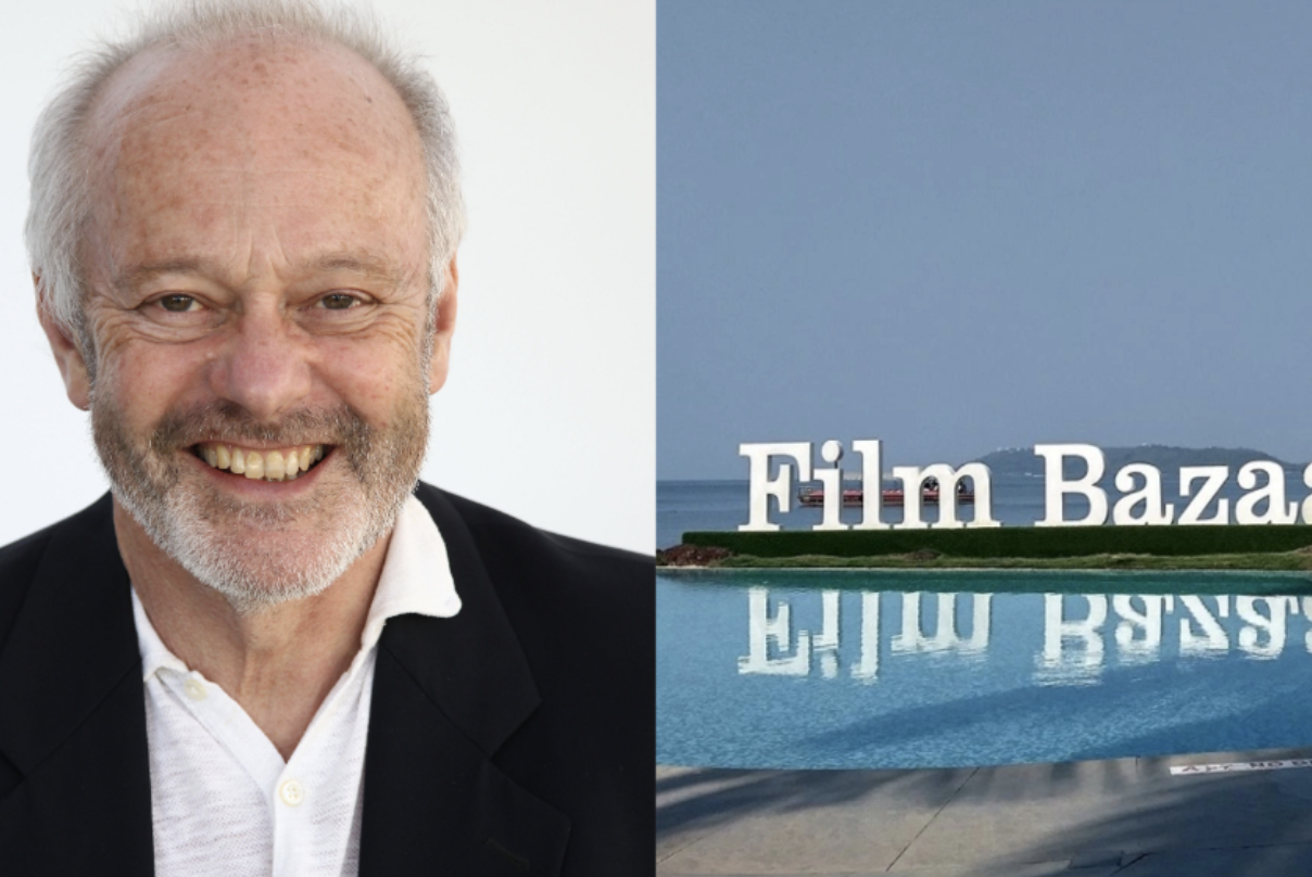 A smiling elderly man with white hair and a beard, wearing a black blazer and white shirt, standing next to a sign that reads 'Film Bazaar' with a background of a beach, water, and a lighthouse.