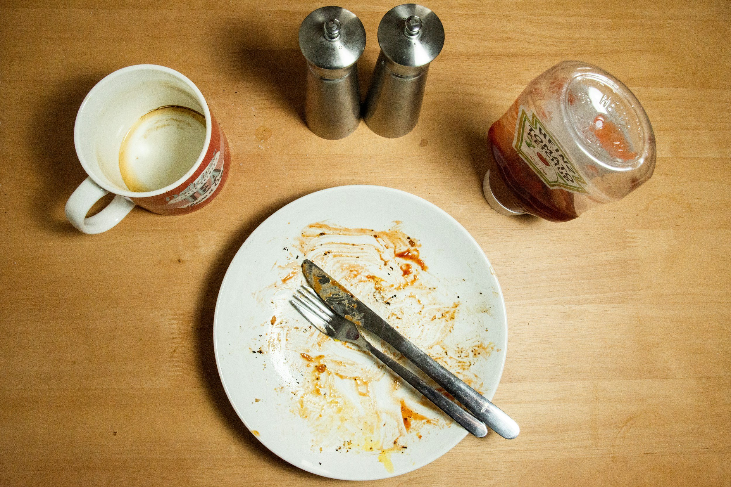An empty plate with cutlery on top an empty mug salt pepper and a empty bottle of tomato ketchup on a brown table 