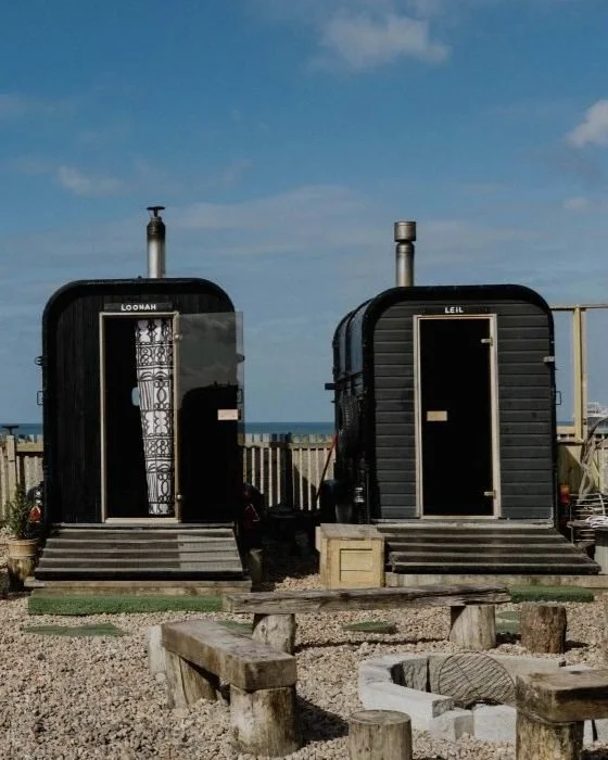 Two small black sauna cabins labeled 'Loonah' and 'Leil' outdoors with steps, in front of a wooden fence and a view of the ocean and sky