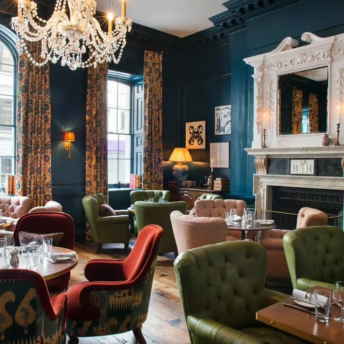 Elegant restaurant interior with dark blue walls, ornate white fireplace, large chandelier, and colorful plush chairs around tables.