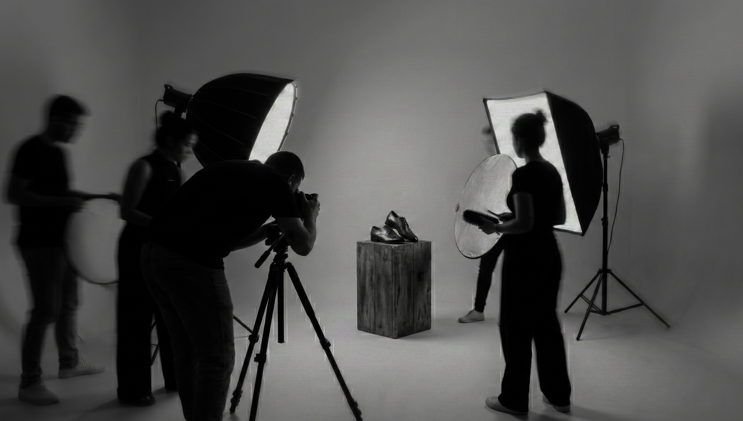 Black-and-white photo of a photo shoot setup with crew, models, and lighting equipment. A pair of shoes on a pedestal in the center.