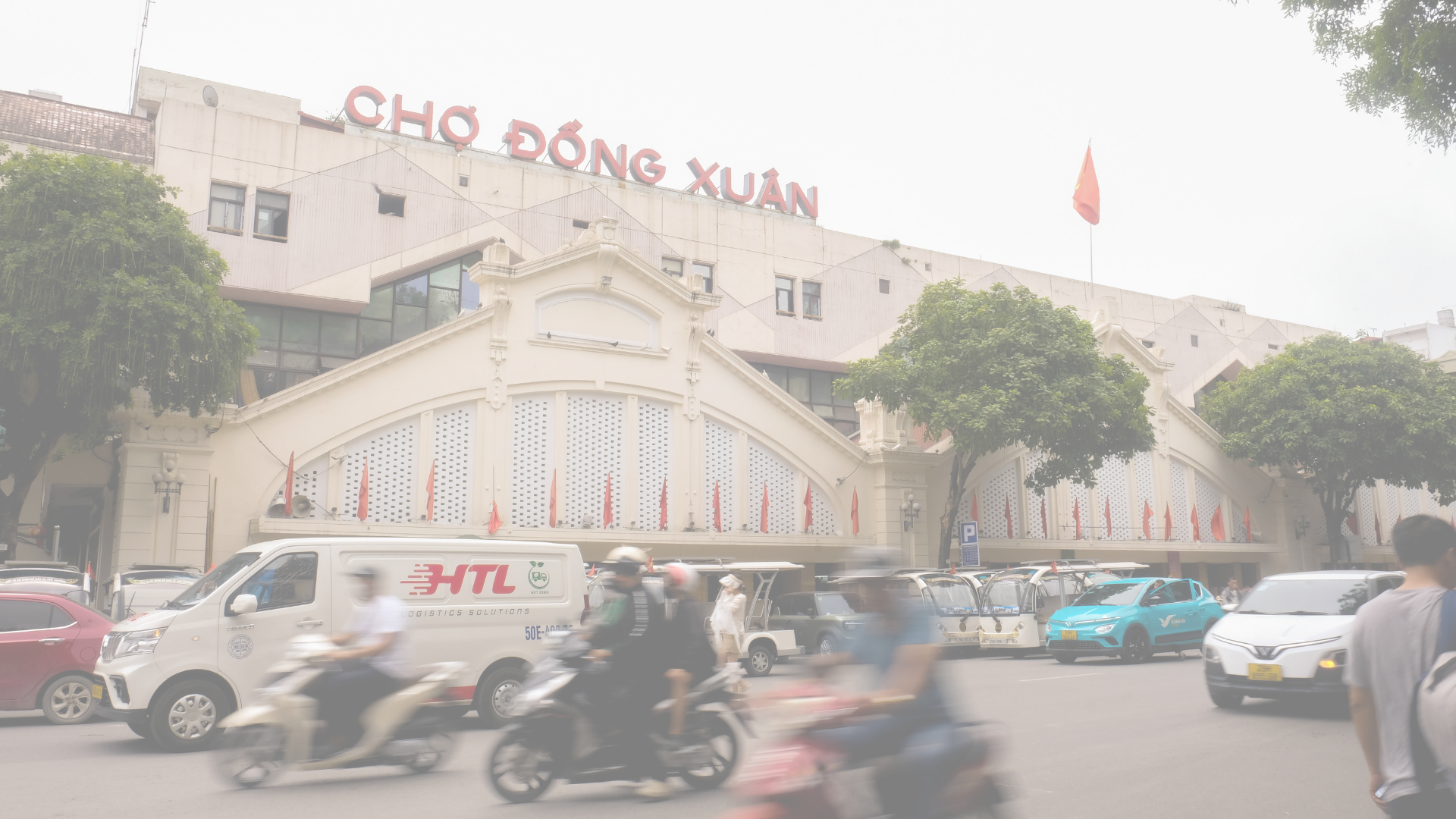 Street scene in front of a large building with a sign in Vietnamese, surrounded by crowded traffic including motorcycles, cars, and a delivery van, with trees and flags visible.