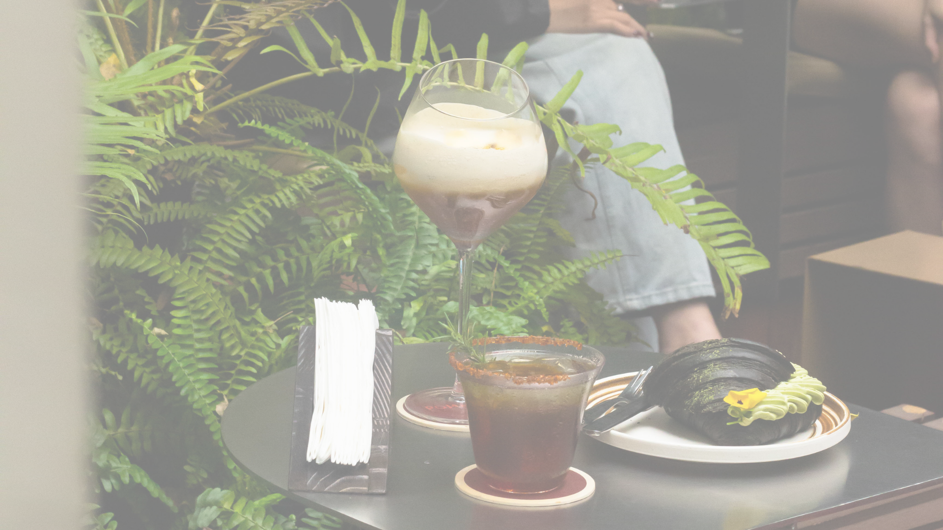 A table with a dessert drink topped with whipped cream, a cocktail glass with a dark beverage, a plate with a black sushi roll decorated with green and yellow flowers, a small bowl of soy sauce, a white napkin, and utensils, with green foliage in the background.