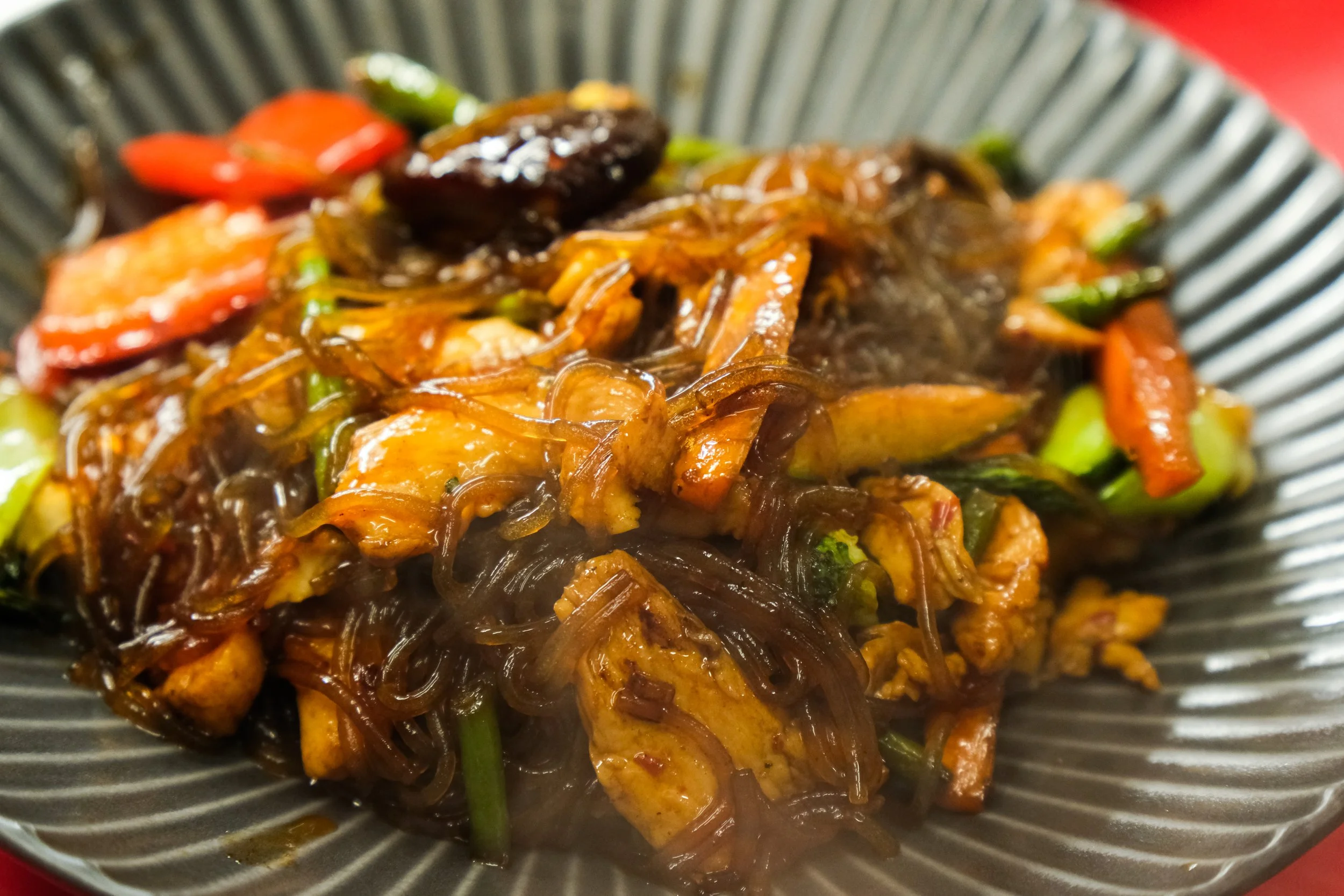 A close-up of stir-fried noodles with vegetables and chicken in a black bowl.