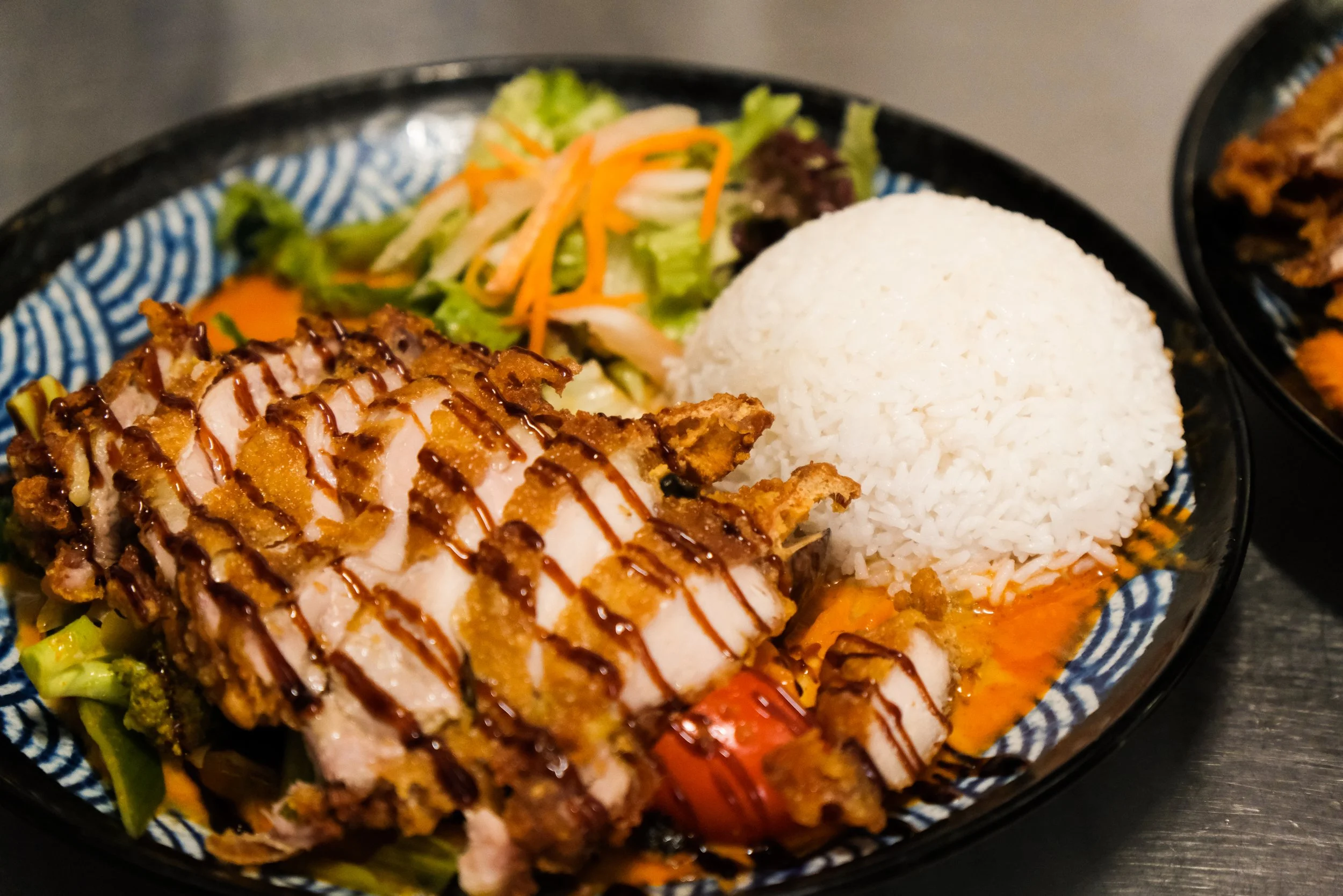 Close-up of a plate with sliced fried chicken topped with sauce, white rice, and a side salad with lettuce, shredded carrots, and cabbage