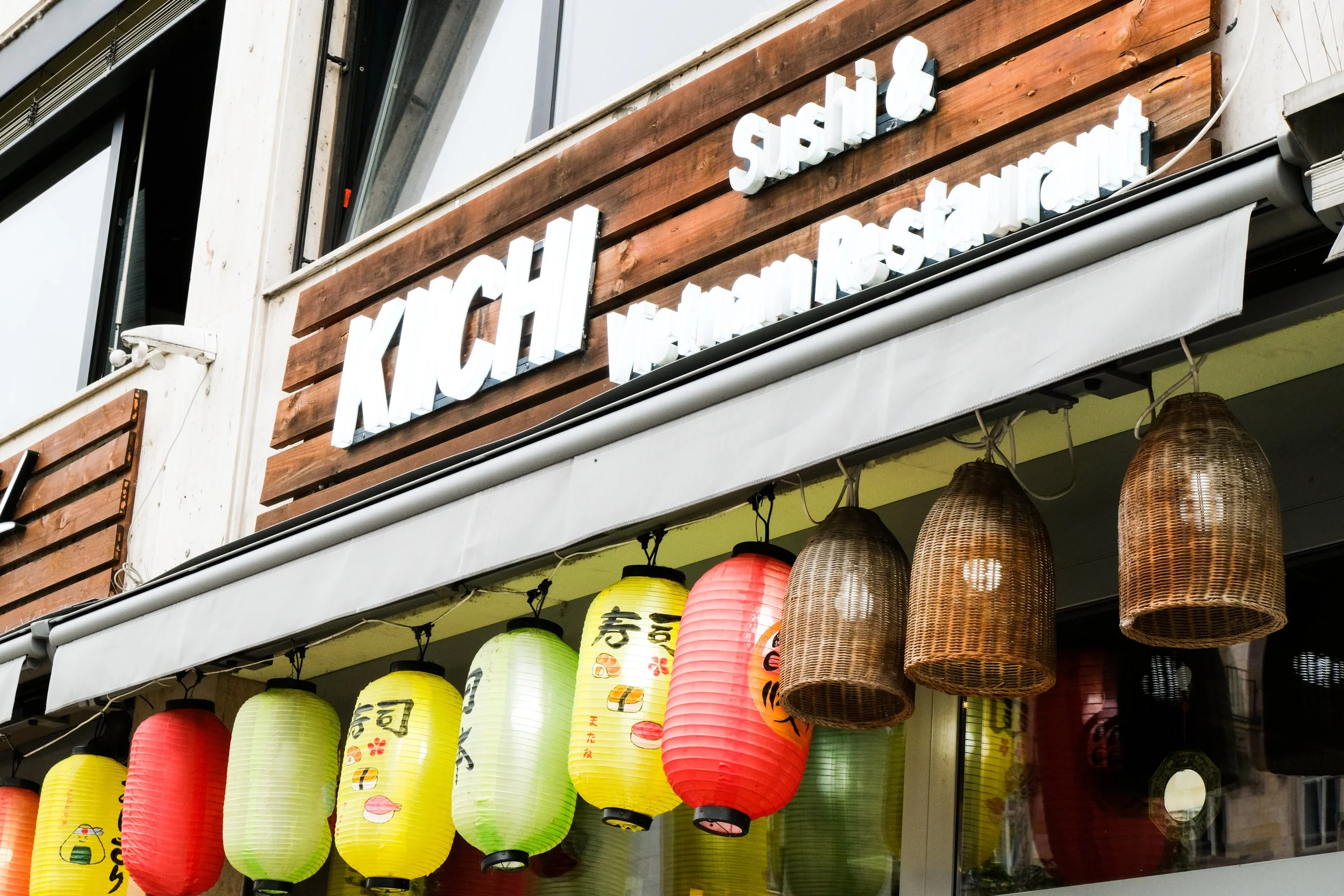 Outdoor view of a restaurant with a wooden sign that says 'KICHI' and smaller words for 'Sushi' and 'Western Restaurant'. Hanging paper lanterns in yellow, red, and green with Japanese characters and designs.