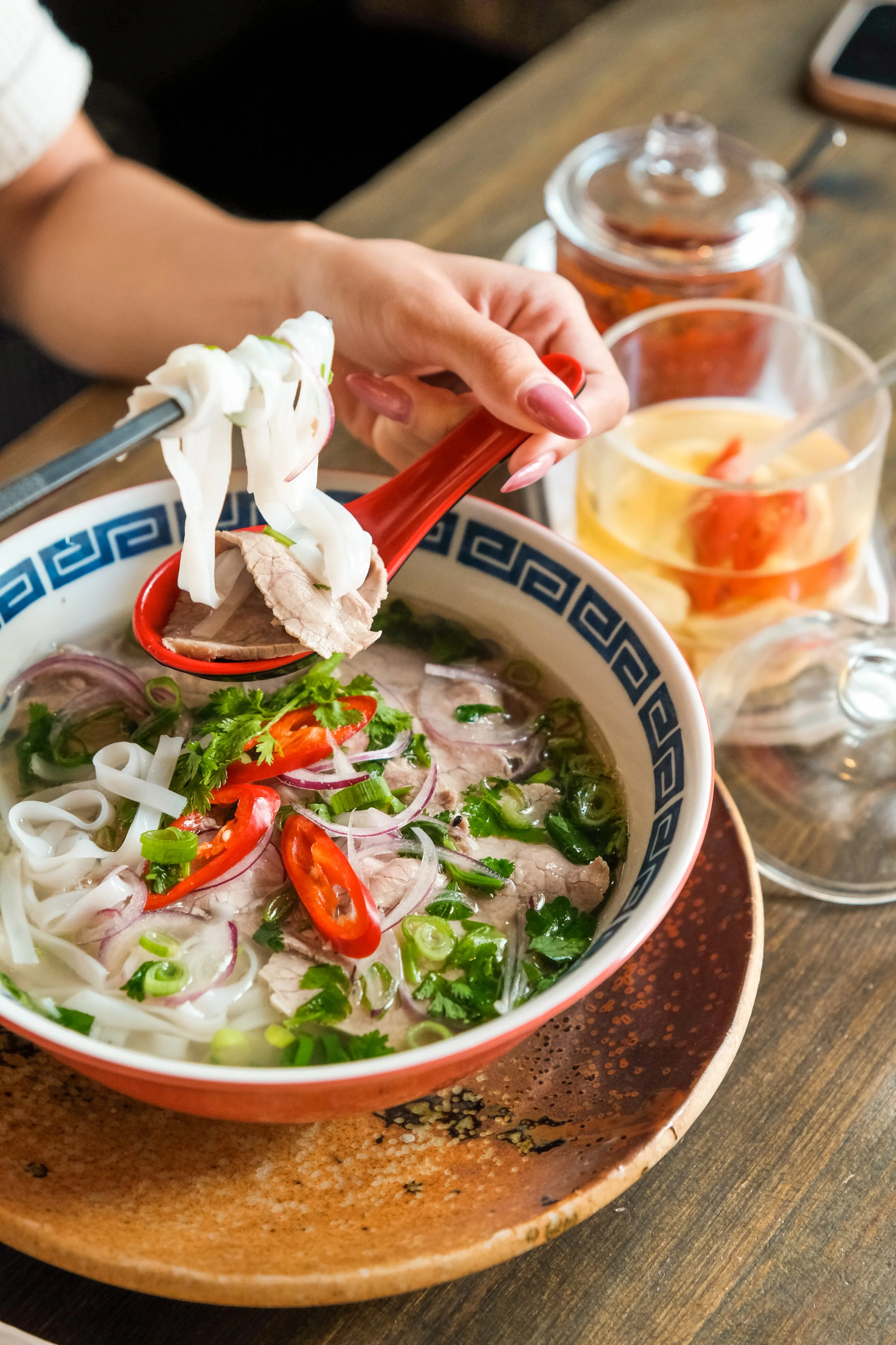 A bowl of Vietnamese pho with slices of beef, rice noodles, red chili peppers, green onions, cilantro, and herbs, with a hand using chopsticks to add more beef. There are small jars containing condiments and lime on the wooden table.