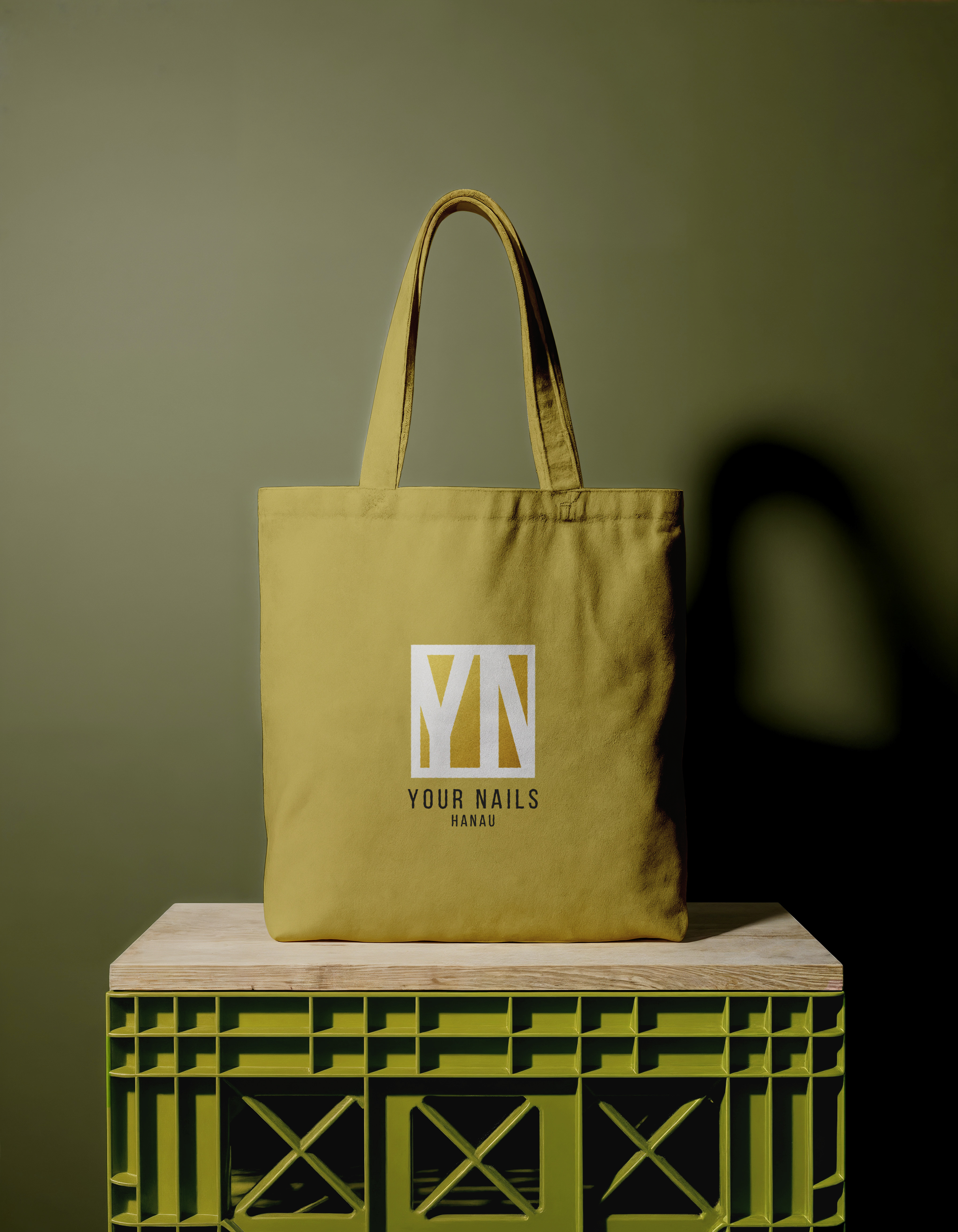 A yellow tote bag with the logo "YN" and the text "Your Nails Hanau" is placed on a wooden surface. The background is a plain wall, and the bag casts a shadow on the wall.