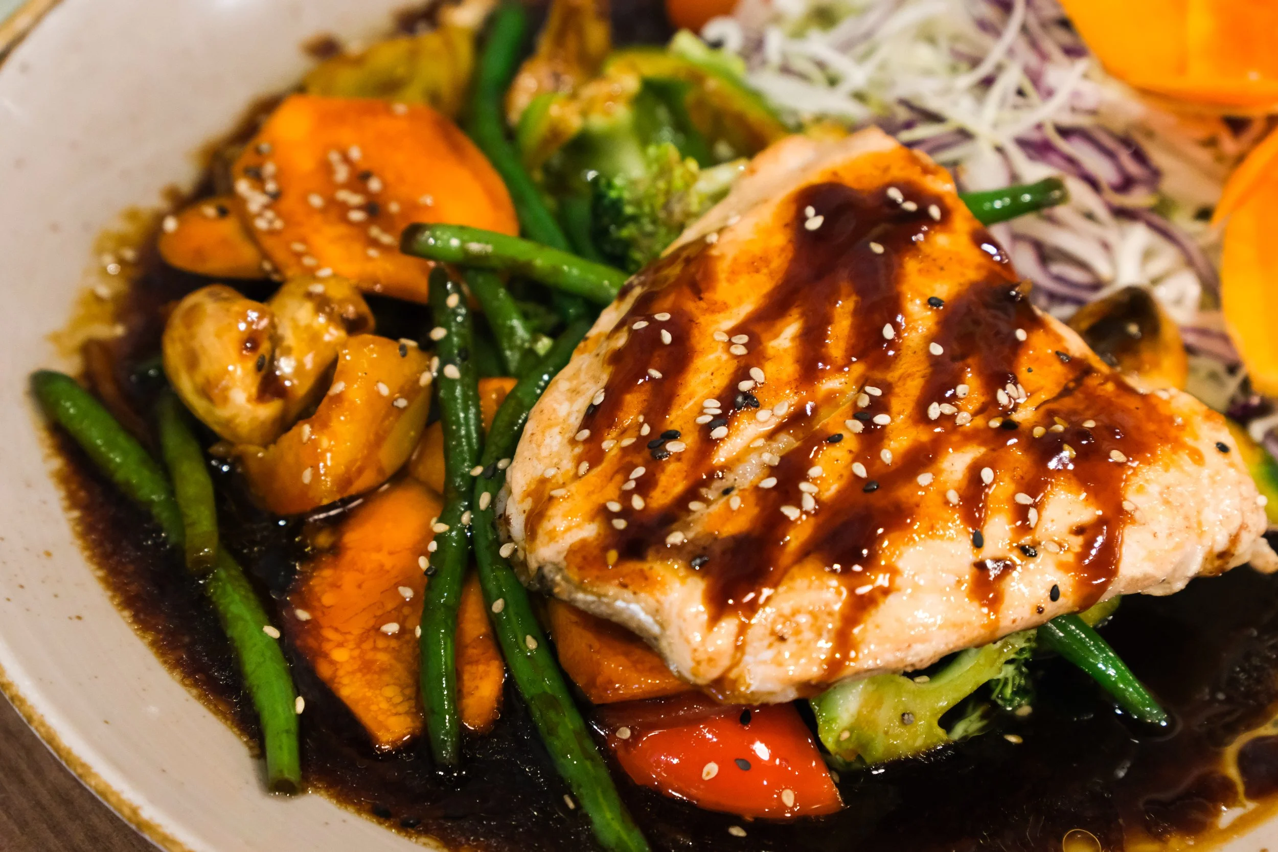 A plate of Asian-inspired cuisine featuring a grilled chicken thigh with dark sauce and sesame seeds, surrounded by sautéed broccoli, green beans, carrots, mushrooms, and sliced yellow bell peppers.