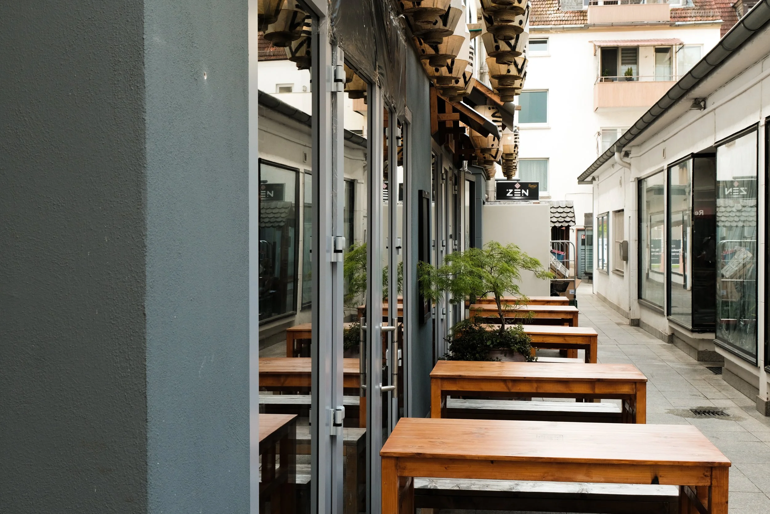 Outdoor seating area with wooden tables and small green trees, located alongside a building with large windows.