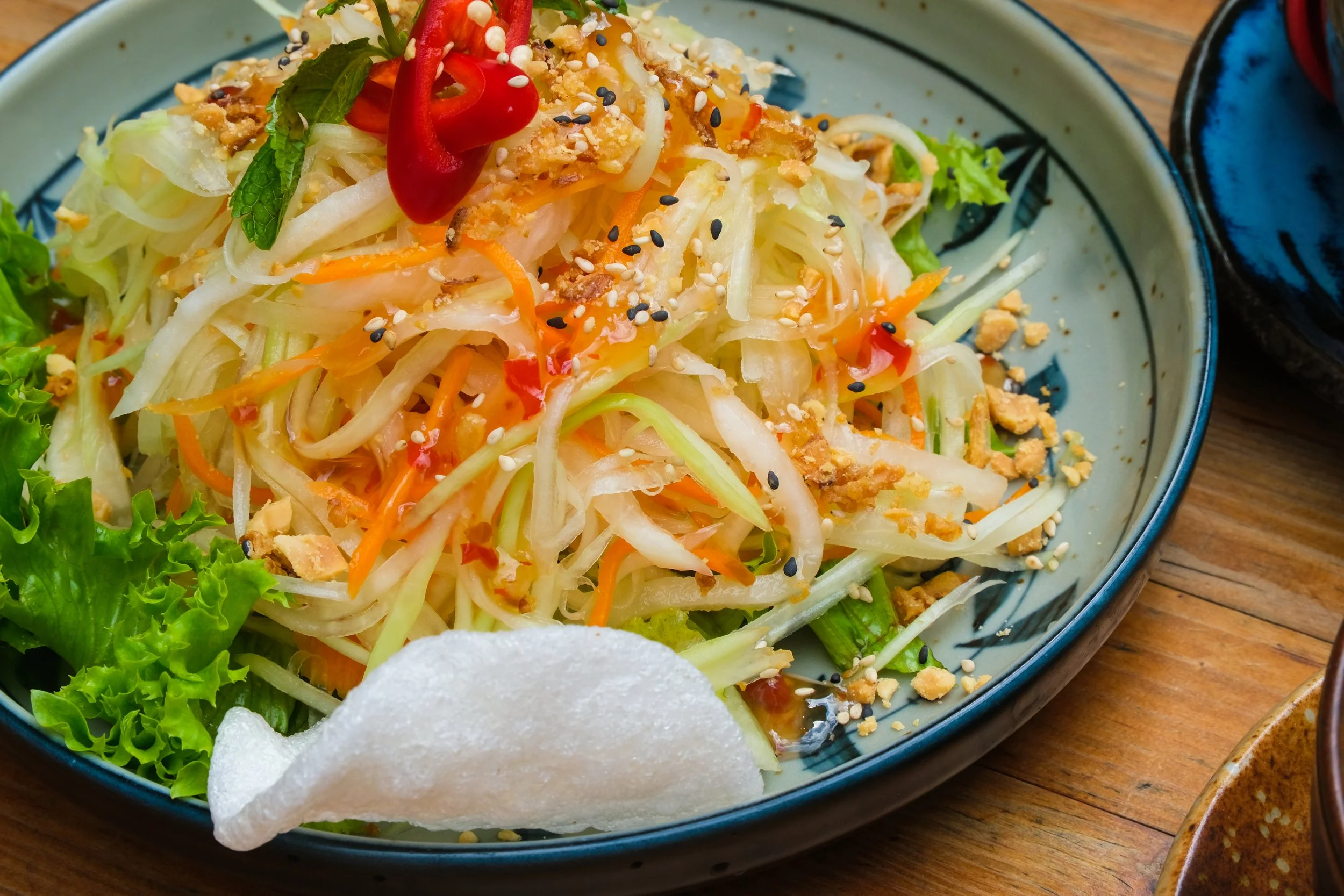Vegetable salad with shredded cabbage, carrots, and lettuce, garnished with red chili, mint leaves, chopped peanuts, sesame seeds, and crispy pork rinds, served in a blue bowl on a wooden table.