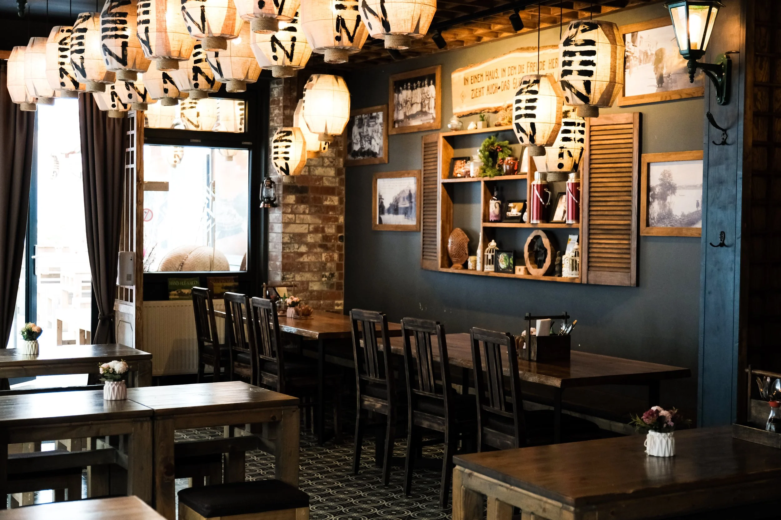 Interior of a cozy restaurant with wooden tables, black chairs, hanging lanterns, and decorative wall art.