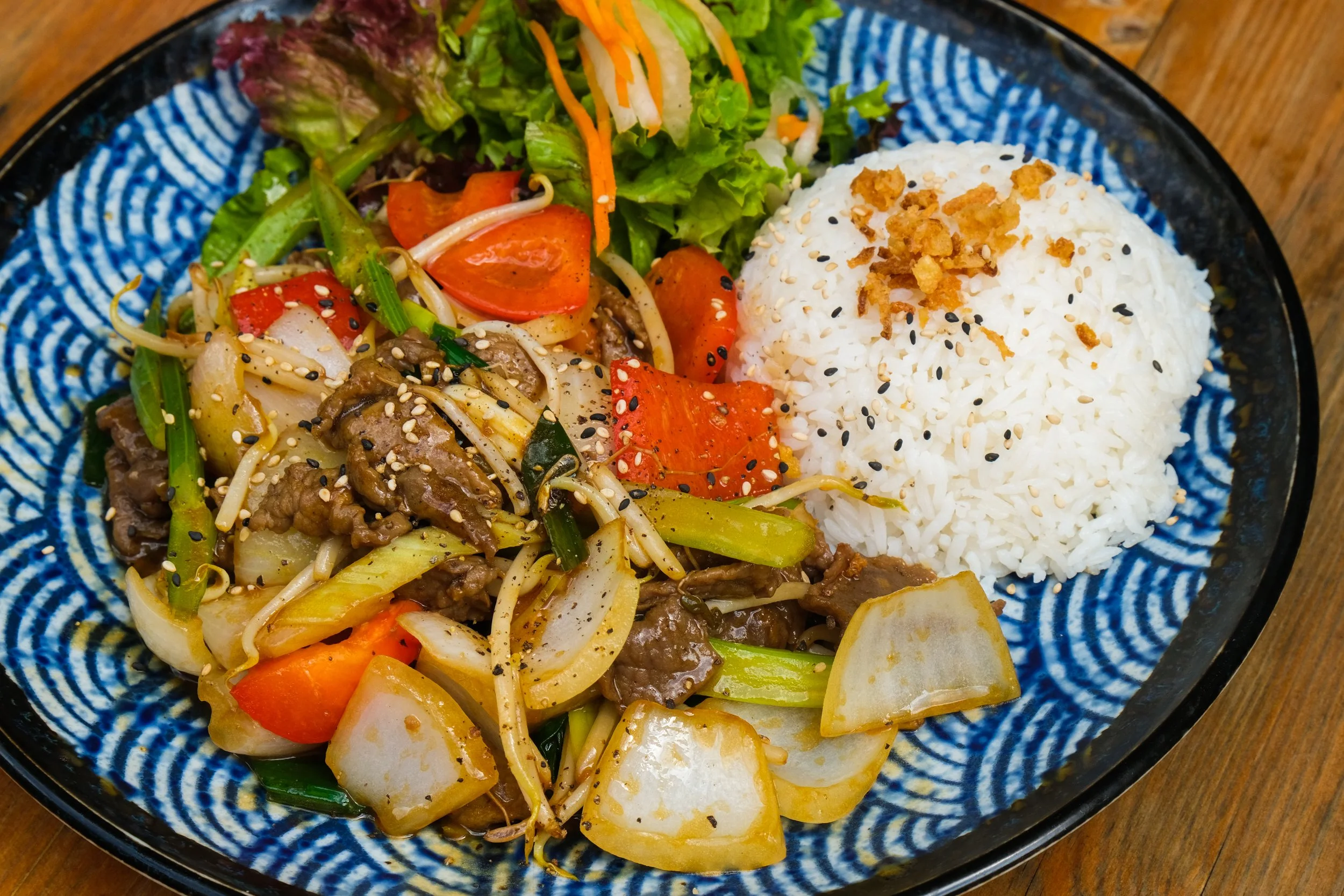 Stir-fry vegetables with beef served with white rice topped with crispy onions, sesame seeds, and black sesame seeds on a decorative blue plate.