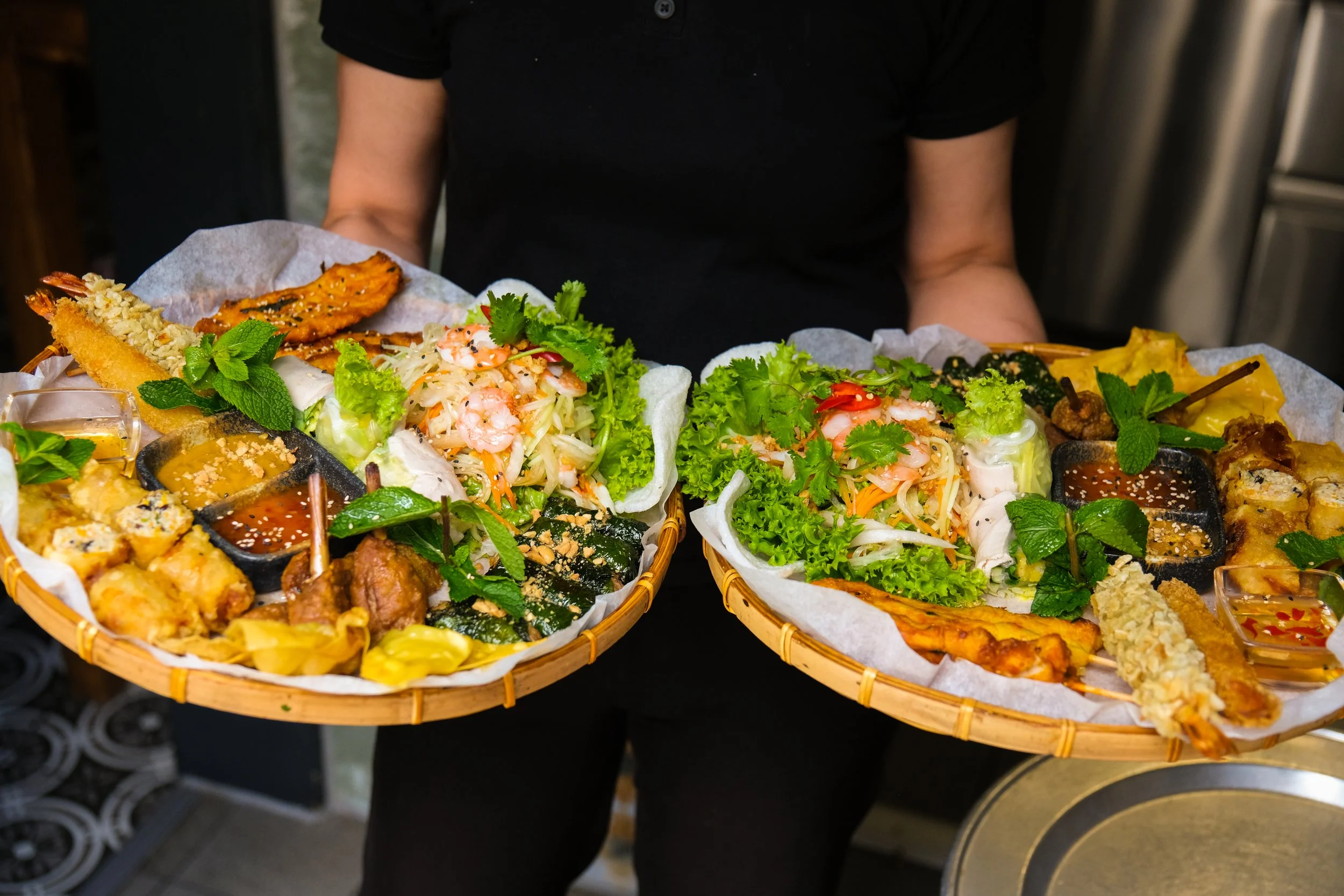 A person holding two baskets filled with assorted Asian cuisine, including vegetables, salads, spring rolls, sauces, and garnishes, in a restaurant setting.