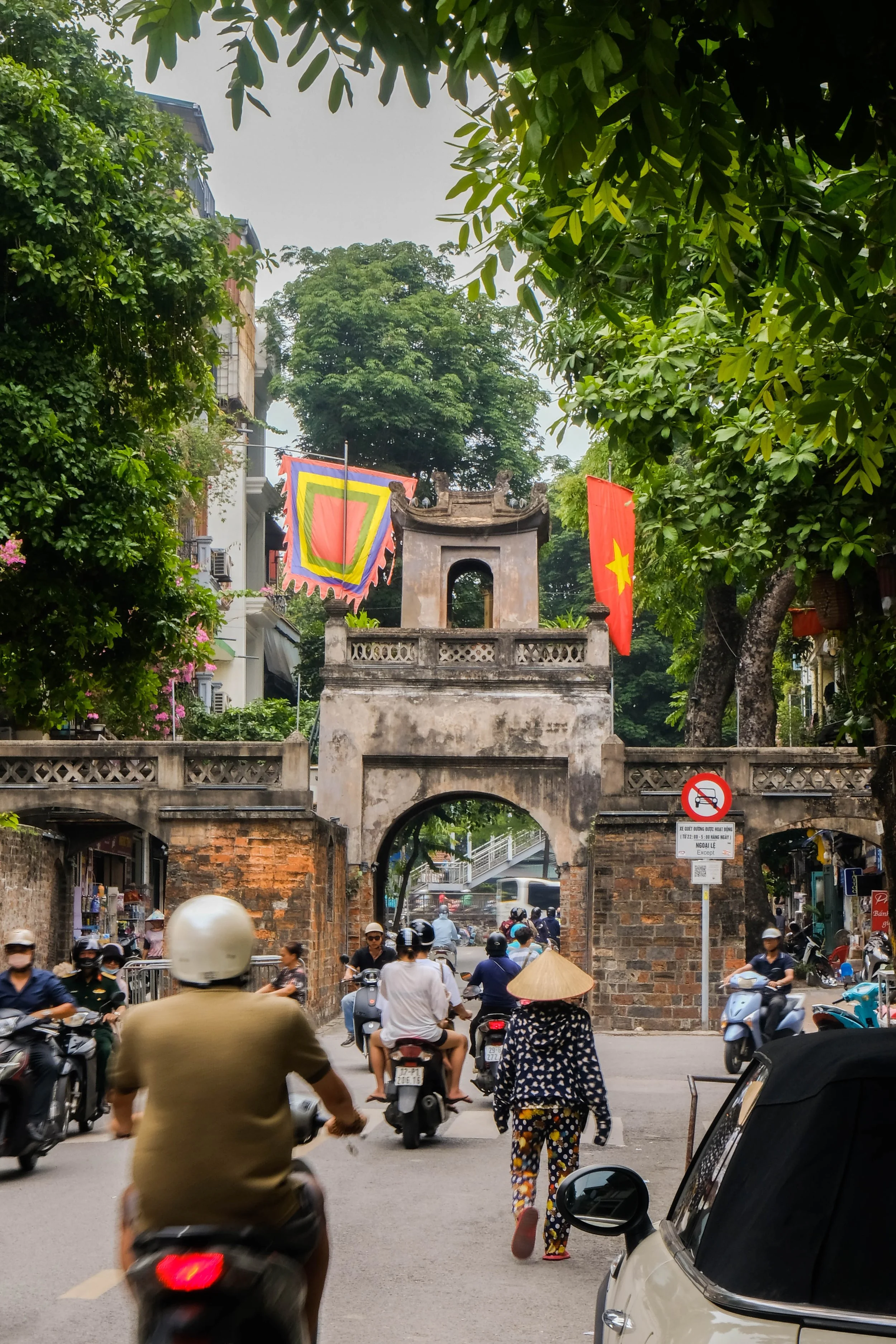 A busy street scene in Vietnam with many motorbikes and scooters, pedestrians, and an old stone archway with flags hanging from it. Lush green trees surround the area.