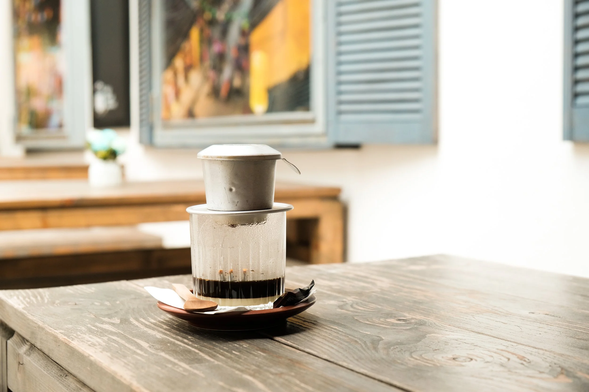 A small metal container sitting on top of a glass filled with dark liquid, placed on a brown ceramic plate on a wooden table.