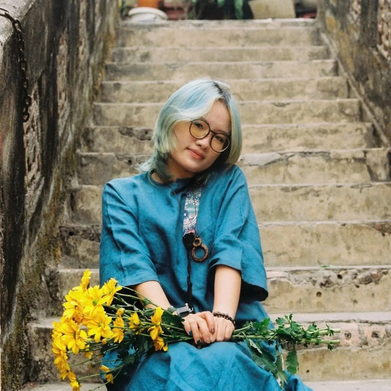 A young woman with dyed blue and blonde hair, wearing glasses and a blue dress, sitting on stone steps outdoors while holding a bouquet of yellow flowers.