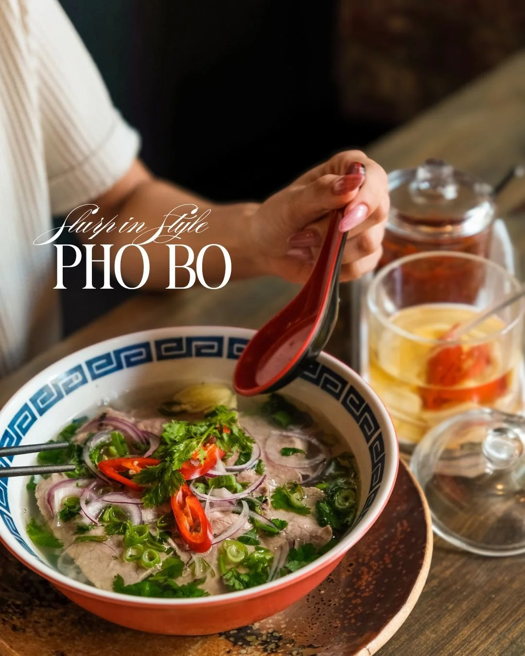 A bowl of pho bo with sliced beef, red chili peppers, red onion, cilantro, and green onions on a wooden table. A person is holding a red spoon over the bowl. There are condiments and a glass of iced tea on the table.