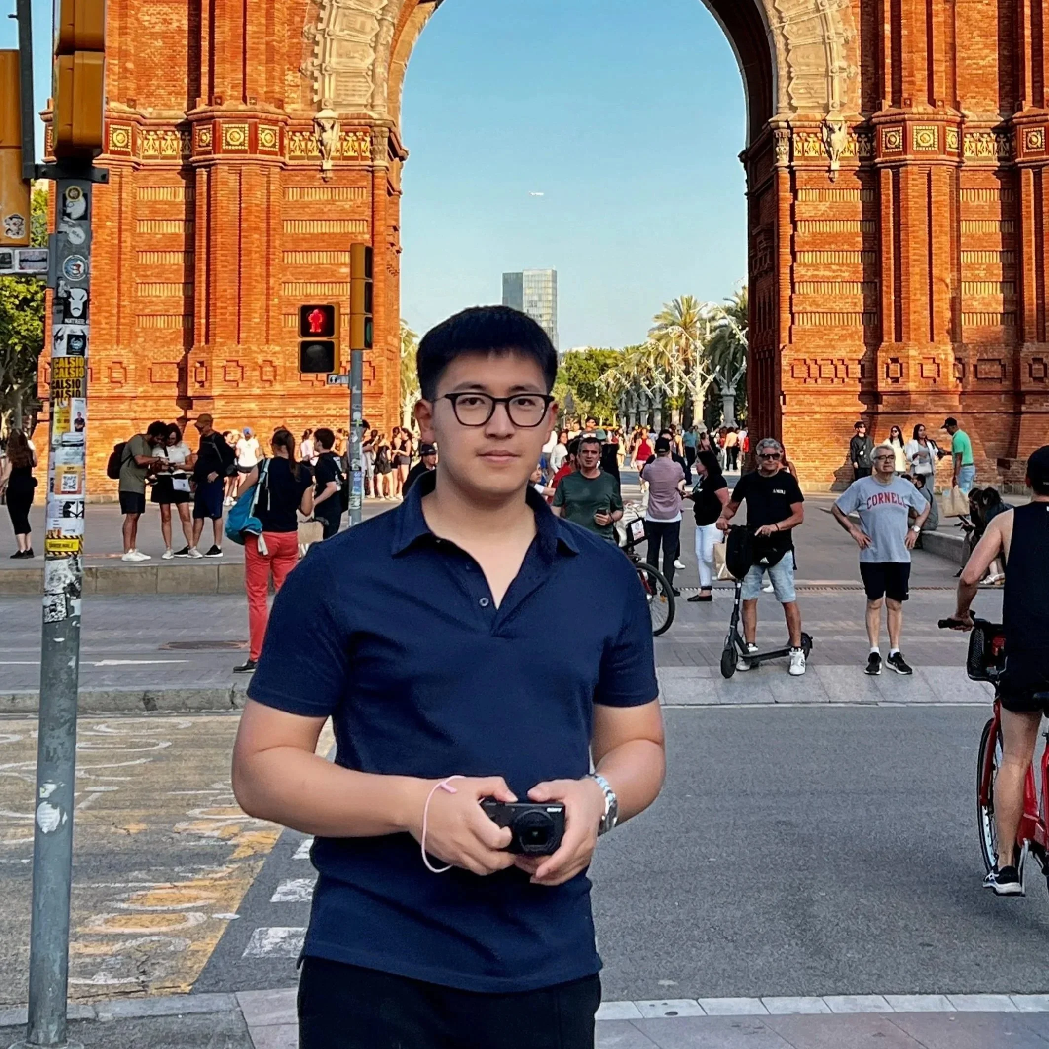 A young man with glasses wearing a navy blue polo shirt holding a camera, standing in front of the historic Arc de Triomphe in a busy city scene, with many people walking and cycling.