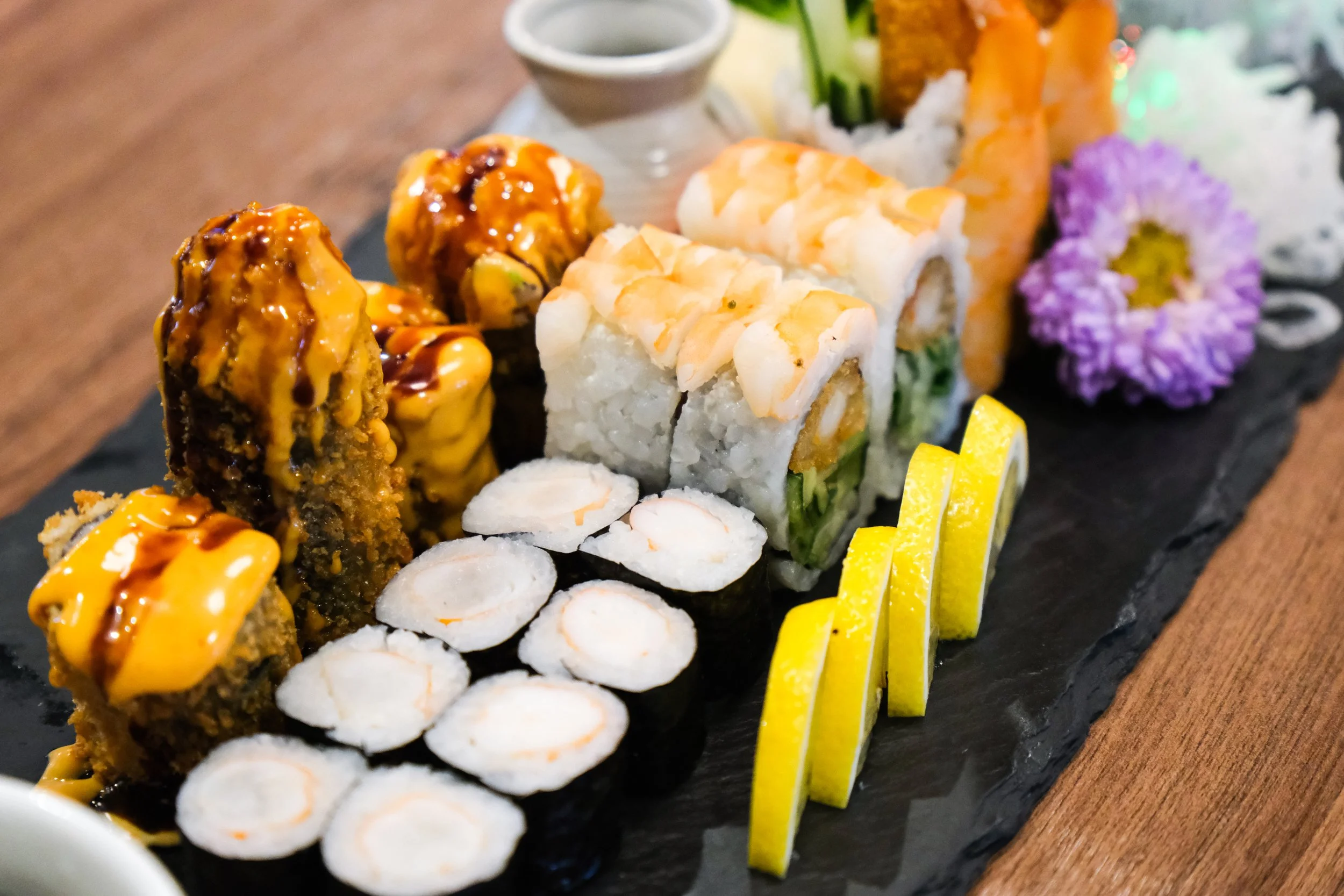 Assorted sushi rolls includes tempura rolls with eel sauce, white fish and avocado rolls with shrimp on top, cucumber and cream cheese rolls, and lemon slices, served on a black slate platter with decorative purple flower.